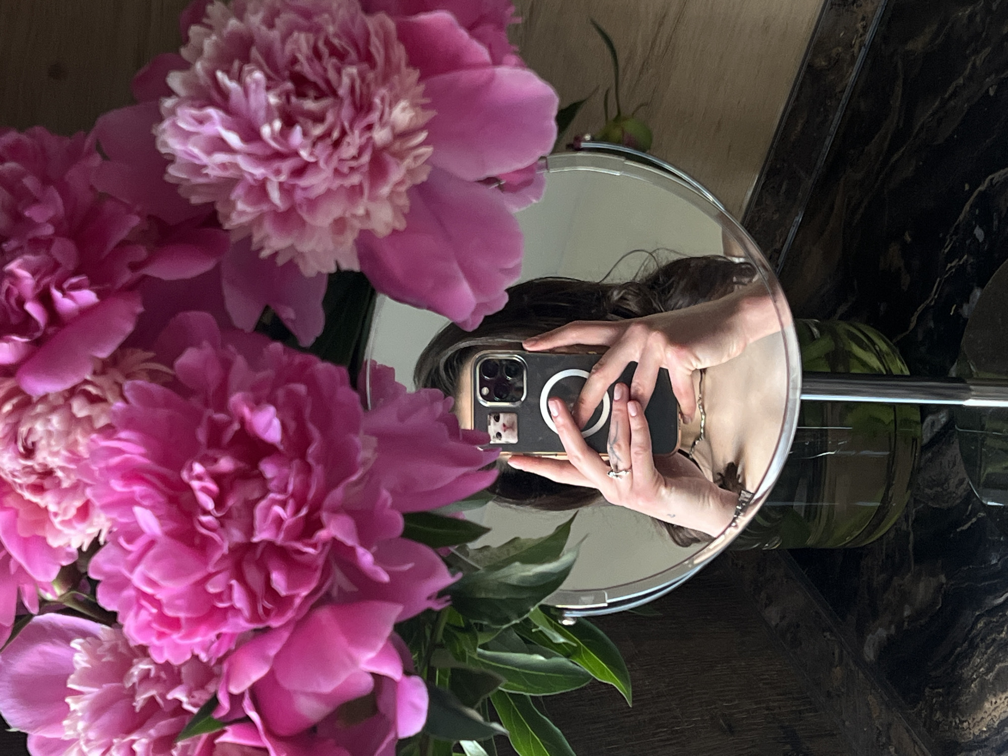A close-up, high-angle, eye-level shot captures a bouquet of bright pink peonies dominating the top half of the frame. The flowers are lush and fully bloomed, with many layers of soft petals. In the lower half of the image, a round mirror reflects a person taking a selfie. The person is holding a smartphone with a decorative sticker of a cat's face on the back. The person is wearing a ring on their left hand and has dark, wavy hair. The reflection also shows a glimpse of the person's shoulder and collarbone. The mirror is mounted on a silver stand, which is visible at the bottom center. The background consists of a dark, textured surface, possibly marble, in the foreground, and a lighter, wood-like surface behind the mirror. The scene appears to be indoors, likely in a domestic setting. The image does not contain any elements that would suggest a specific location in Ternopil, Ukraine.