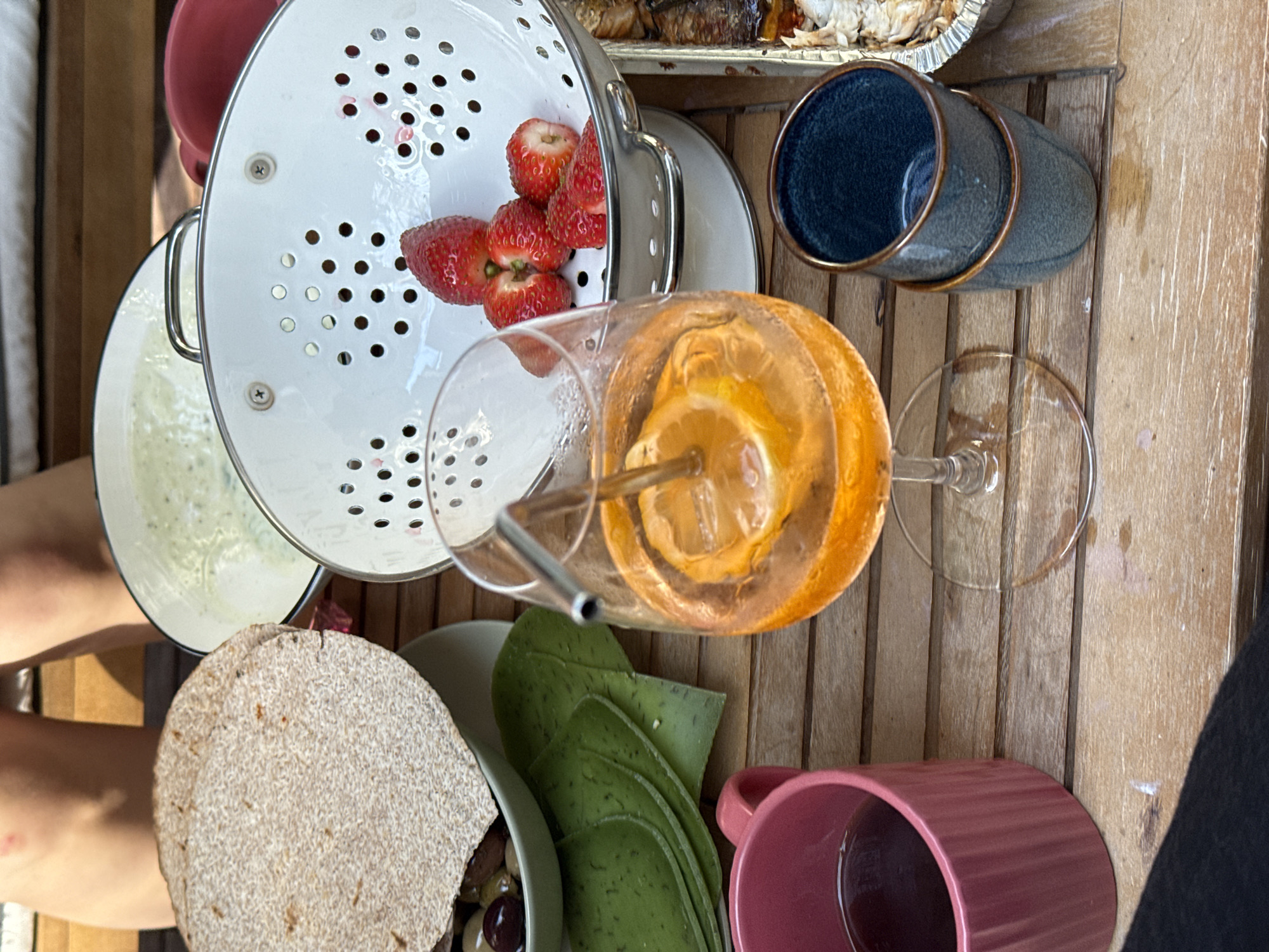 The image shows a table setting, likely outdoors given the wooden planks of the table. The table is covered with various dishes and objects: a white colander with strawberries, a white bowl with a light-colored liquid, a glass of orange-colored drink with a metal straw, a plate of flatbreads and green-colored sliced food, a pink mug, and a couple of blue ceramic cups. The location cannot be determined definitively as Tel-Aviv, Israel. The picture composition strongly suggests a casual meal setup. The image details offer information limited to the mentioned tangible items and their spatial arrangement. Without additional data, elements like specific interactions, the time of day, or precise environmental circumstances remain unknown.
