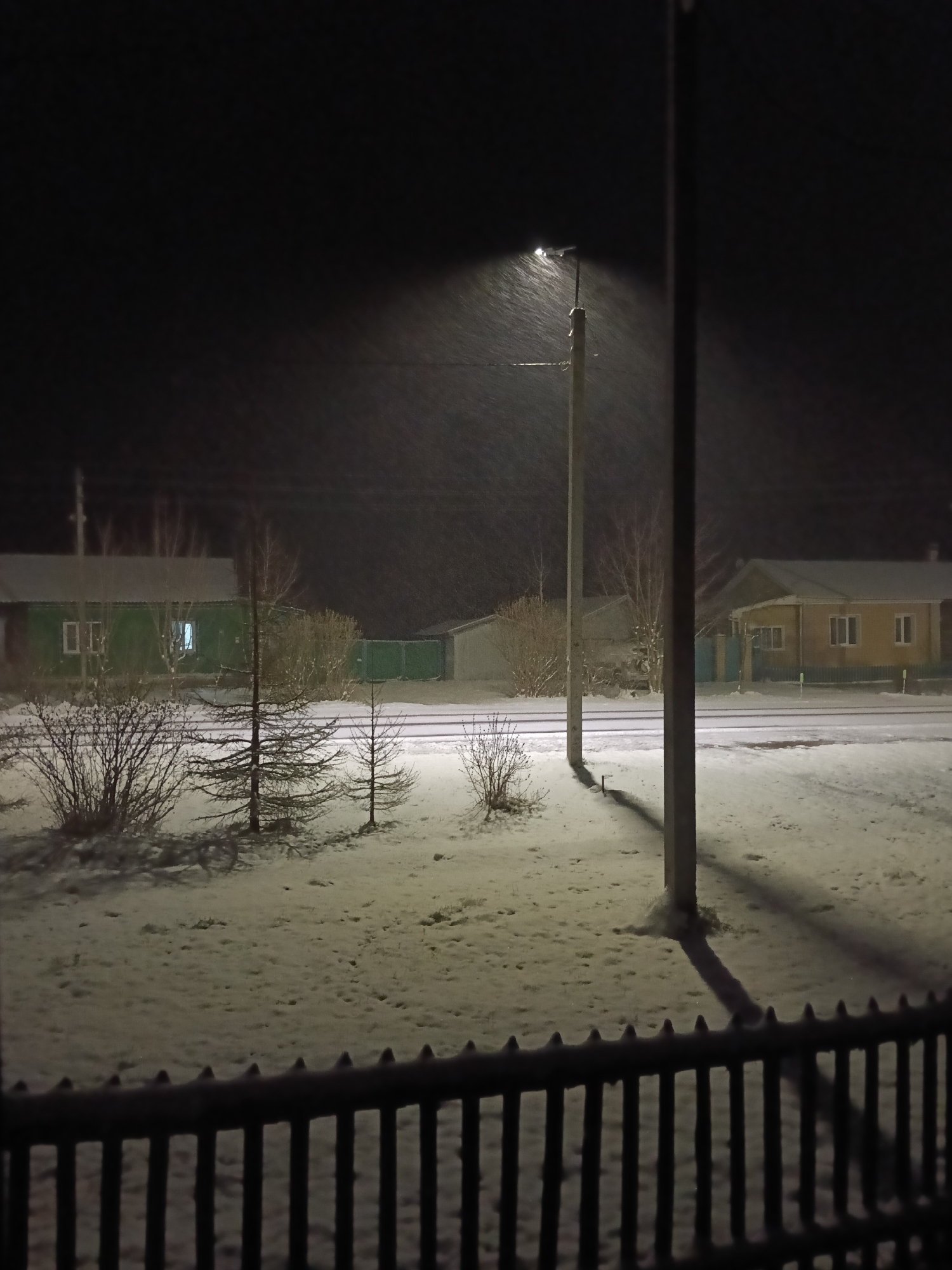 The image depicts a night scene in Красный Маяк, Russia, during a snowfall. The foreground features a dark, wooden picket fence. Beyond the fence is a snow-covered yard with sparse vegetation, including small bushes and young coniferous trees. A street separates the yard from several single-story houses. The houses are illuminated by internal lights, with at least two of them painted in light colors. A streetlamp in the middle ground casts a bright light that illuminates the falling snow. Two utility poles are visible, one lit and the other obscured in darkness. The background is a dark, snow-filled sky.