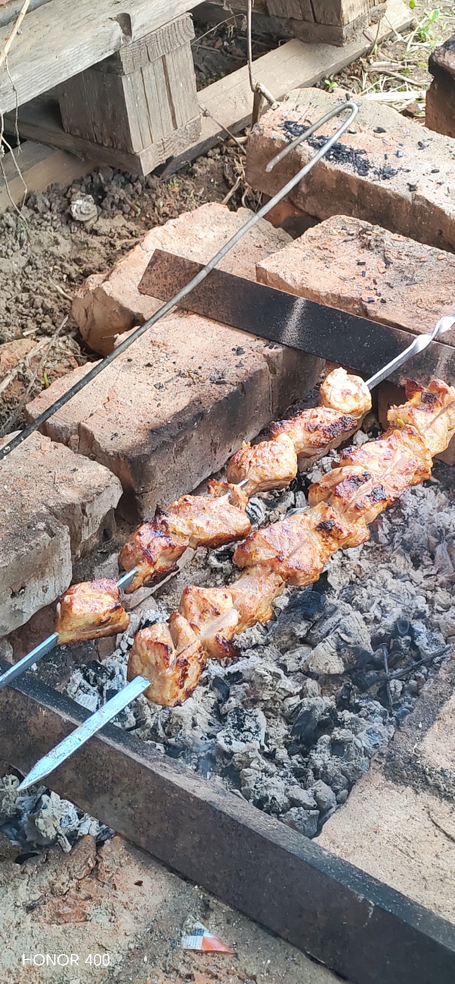 The image shows two skewers with cooked meat, likely shashlik, resting on a makeshift grill constructed from bricks and metal bars. The grill sits above a bed of gray, ashy embers. The surroundings appear to be outdoors, possibly a backyard or open area in Noviy, Russia, indicated by the camera's watermark "HONOR 400". The ground is visible around the grilling area, showing patches of bare earth, some wooden planks and scattered debris. The focus of the image is on the cooked meat, highlighting its browned and slightly charred exterior.