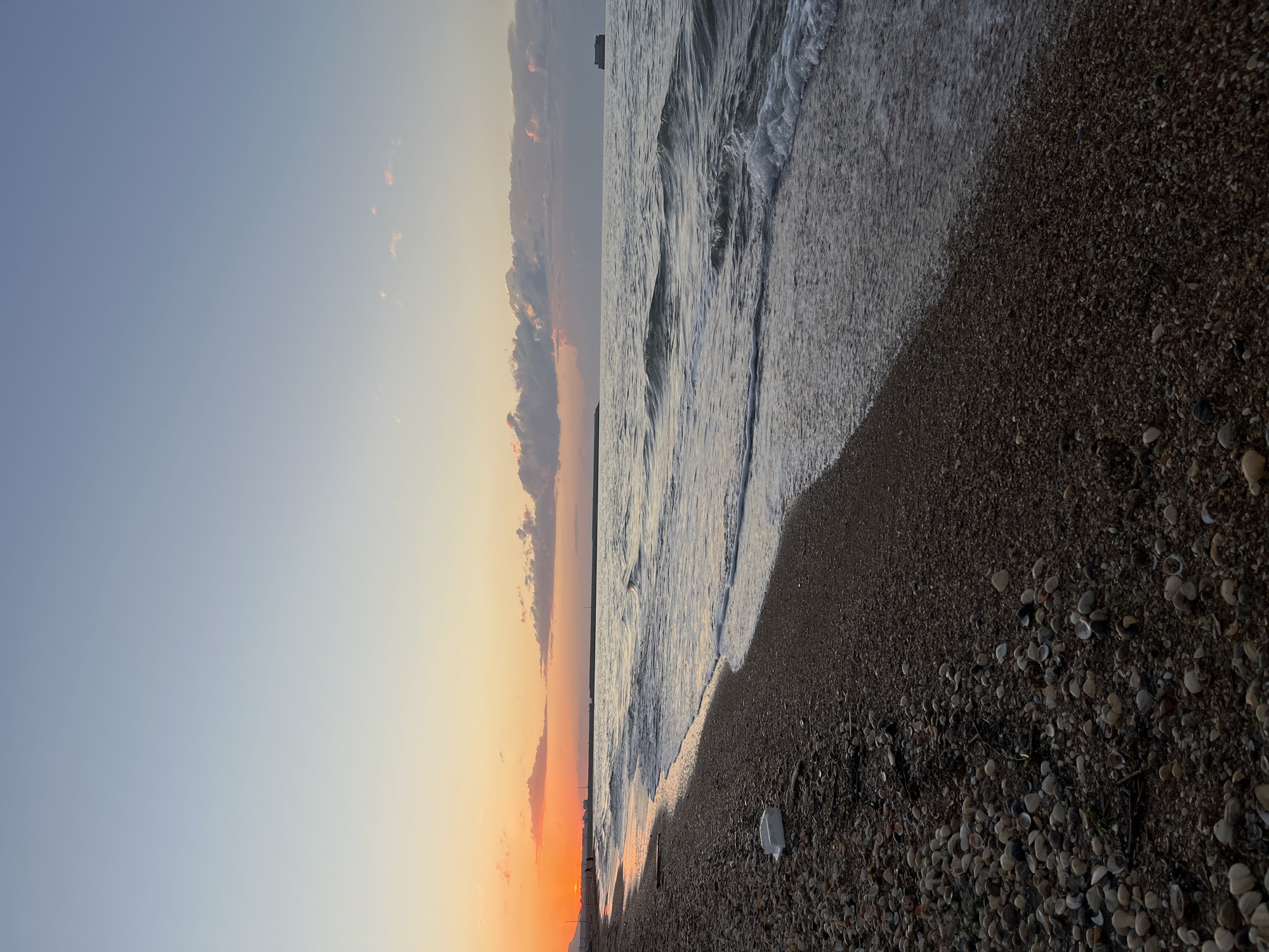 The image depicts a sunset view of the Caspian Sea in Kaspiysk, Russia. The sky transitions from a clear blue to a warm orange near the horizon, with some clouds tinged with pink and orange hues. The sea is moderately choppy with visible waves approaching the shore. The beach is composed of small pebbles and numerous seashells. There is a piece of white trash on the beach. A distant structure, possibly a building or ship, is faintly visible on the horizon. The image is oriented vertically.