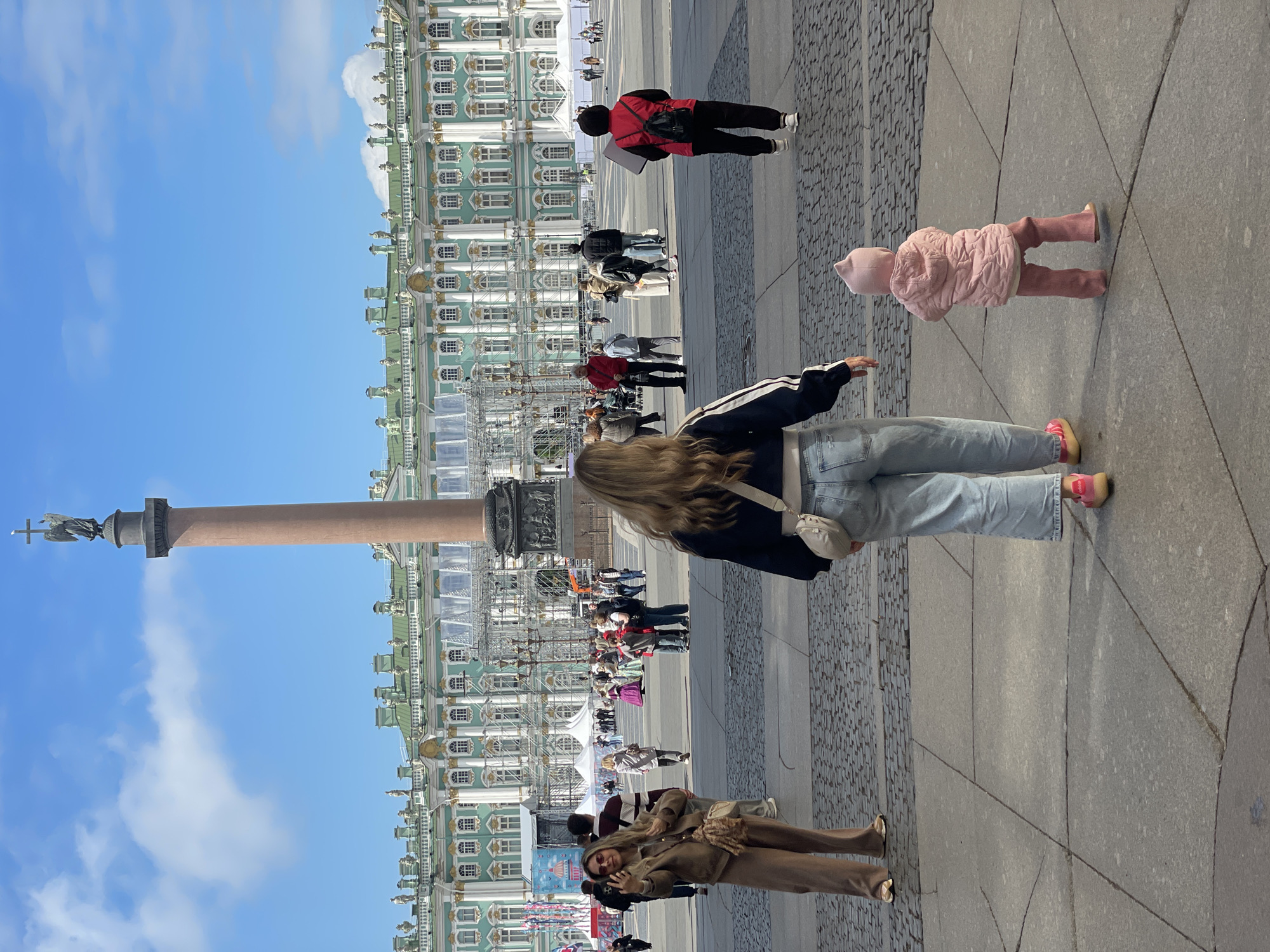The image captures a scene in the Palace Square in Saint Petersburg, Russia. In the background is the Winter Palace, painted in a distinctive blue-green color, with scaffolding erected in front of it. A tall column stands in the square, topped with a statue. Several people are present. A young child, wearing a pink snowsuit, stands near a woman dressed in jeans and a black and white jacket. Near the bottom of the image, another woman, dressed in a brown suit and wearing sunglasses, makes a peace sign with her hand. Other people are walking in the background, some appearing to be tourists. The sky is bright blue with scattered clouds. The ground is covered in a mix of concrete and cobblestone.