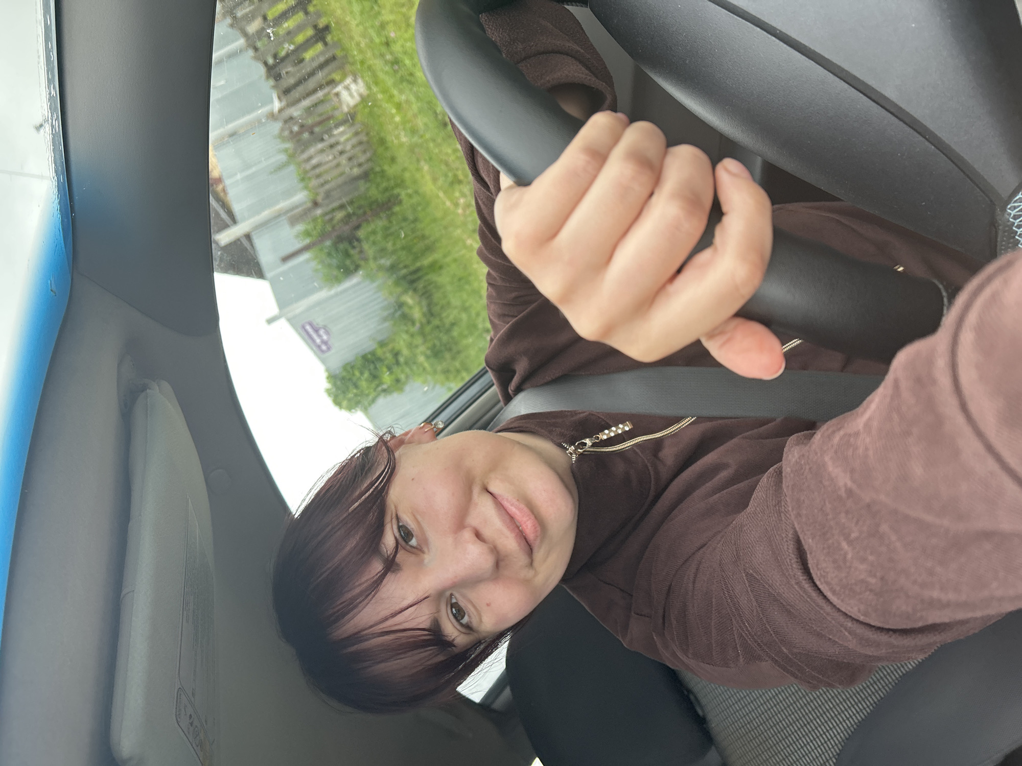 The image shows a young woman with dark hair and bangs, wearing a brown zip-up hoodie. She is inside a car, holding the steering wheel with her right hand. Her left hand is also visible, resting on the steering wheel. The woman is smiling and looking at the camera, which is positioned to take a selfie.
The background visible through the car window suggests an outdoor setting with green foliage and some wooden structures, possibly a fence or a building. The lighting indicates it's daytime, with overcast or diffused light suggesting it might be a cloudy day. The overall mood appears relaxed and cheerful.
There is no visible text in the image. The scene is set within a vehicle, and the activity is a woman taking a selfie while in the driver's seat. The specific location is identified as Фоки, Russia.