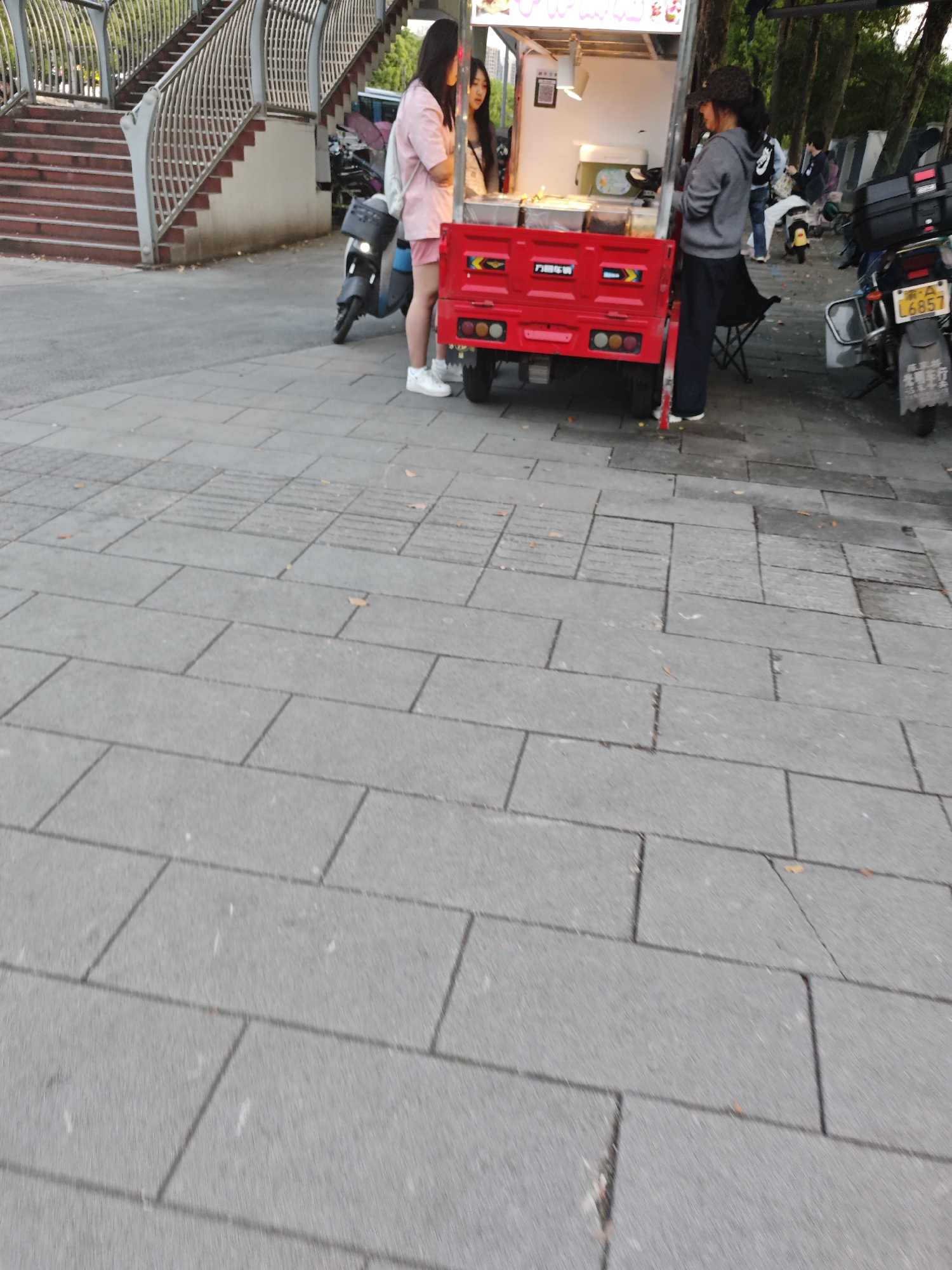 The image, taken in the Chongqing High-tech Zone, China, shows a street scene. A red, three-wheeled vehicle with a food stall attached is the central focus. Two women are standing at the stall. One woman, dressed in pink shorts and a top, is standing in front of the stall. The second woman is standing behind the stall. A third person, wearing a gray jacket and dark pants, stands to the right of the stall. Another parked scooter with a license plate number is visible behind the stall. The background includes a staircase leading to a pedestrian walkway. The ground is paved with large gray rectangular blocks. The setting suggests a public area where food vendors operate.