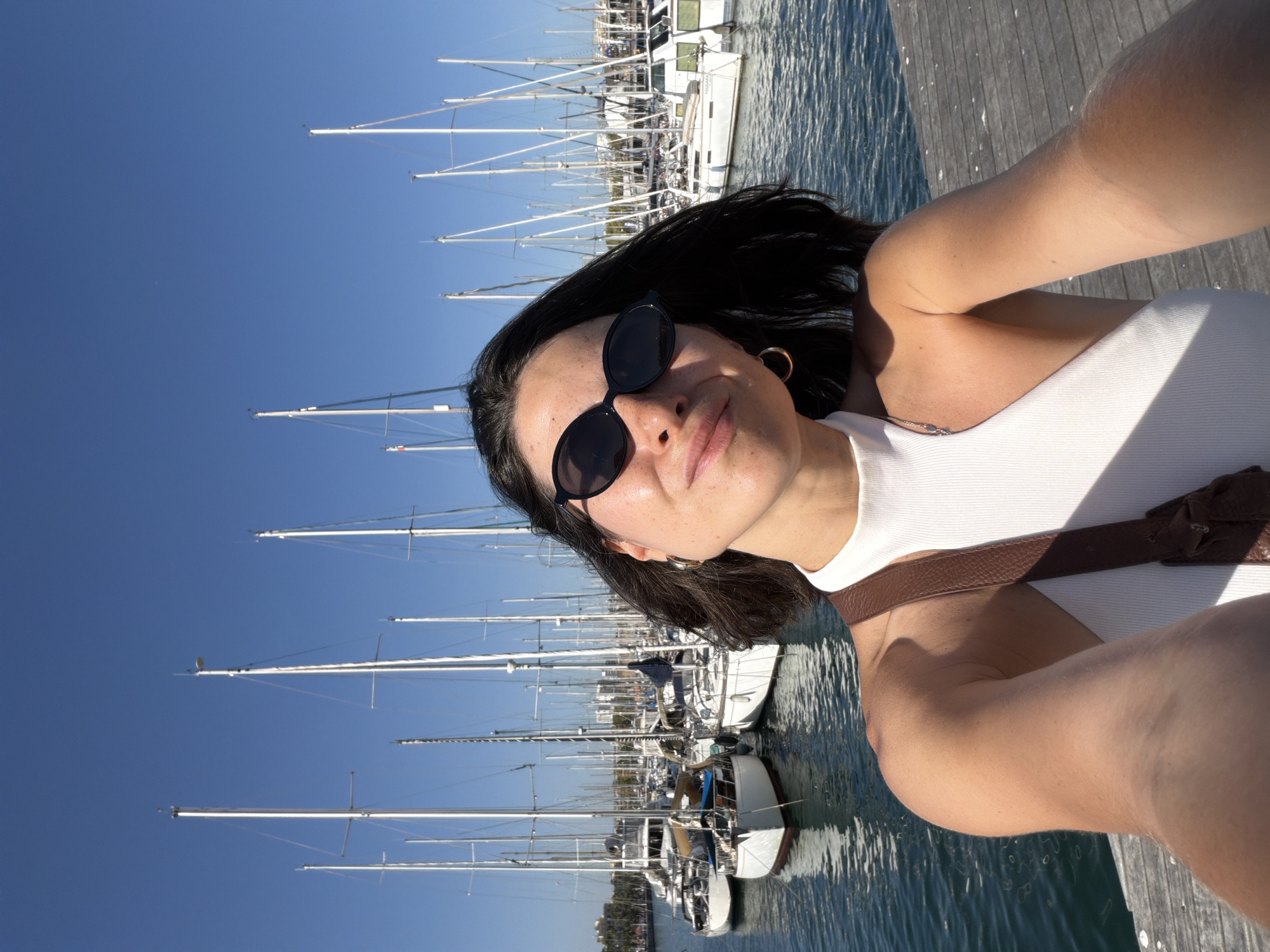 A woman is taking a selfie with a marina full of sailboats in the background. She is wearing a white halter top and black sunglasses. The water is calm and the sky is clear and blue. The marina appears to be located in Barcelona, Spain, possibly near the Port Olímpic area given the density of sailboats and the architectural style of the distant buildings.