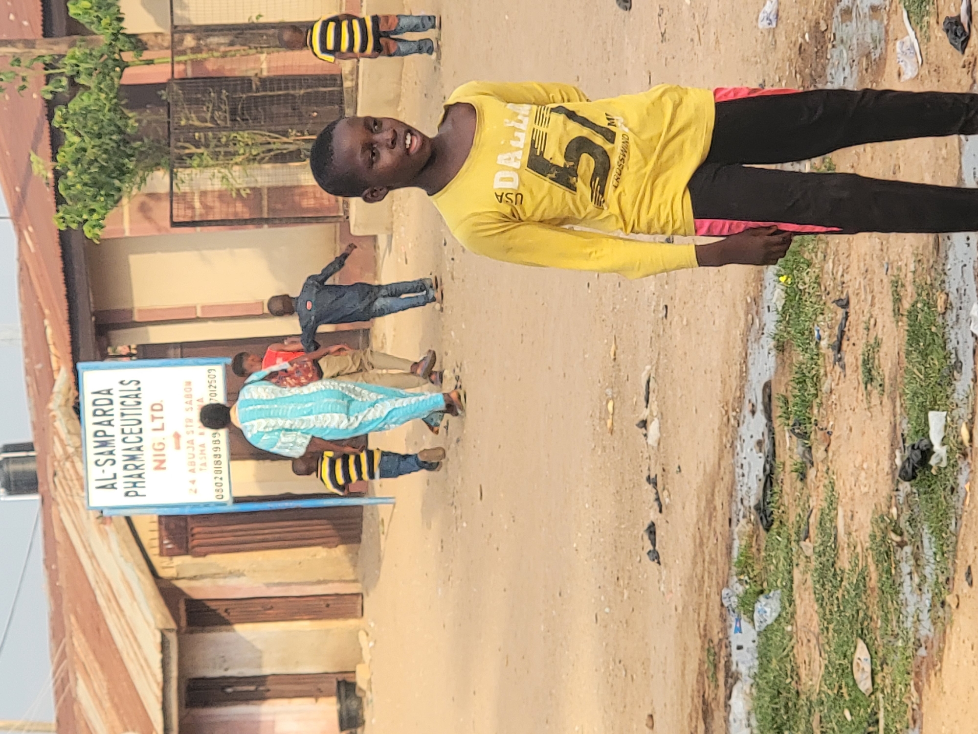 A scene in Sabon Tasha, Nigeria shows a young person wearing a yellow shirt and black pants is the focus of the image. The person is standing on a dirt path. Several other people are in the background, near a building with a sign reading "AL-SAMPARDA PHARMACEUTICALS NIG. LTD." with an address and phone numbers listed. The building appears to be of simple construction. The surrounding environment is littered with trash.