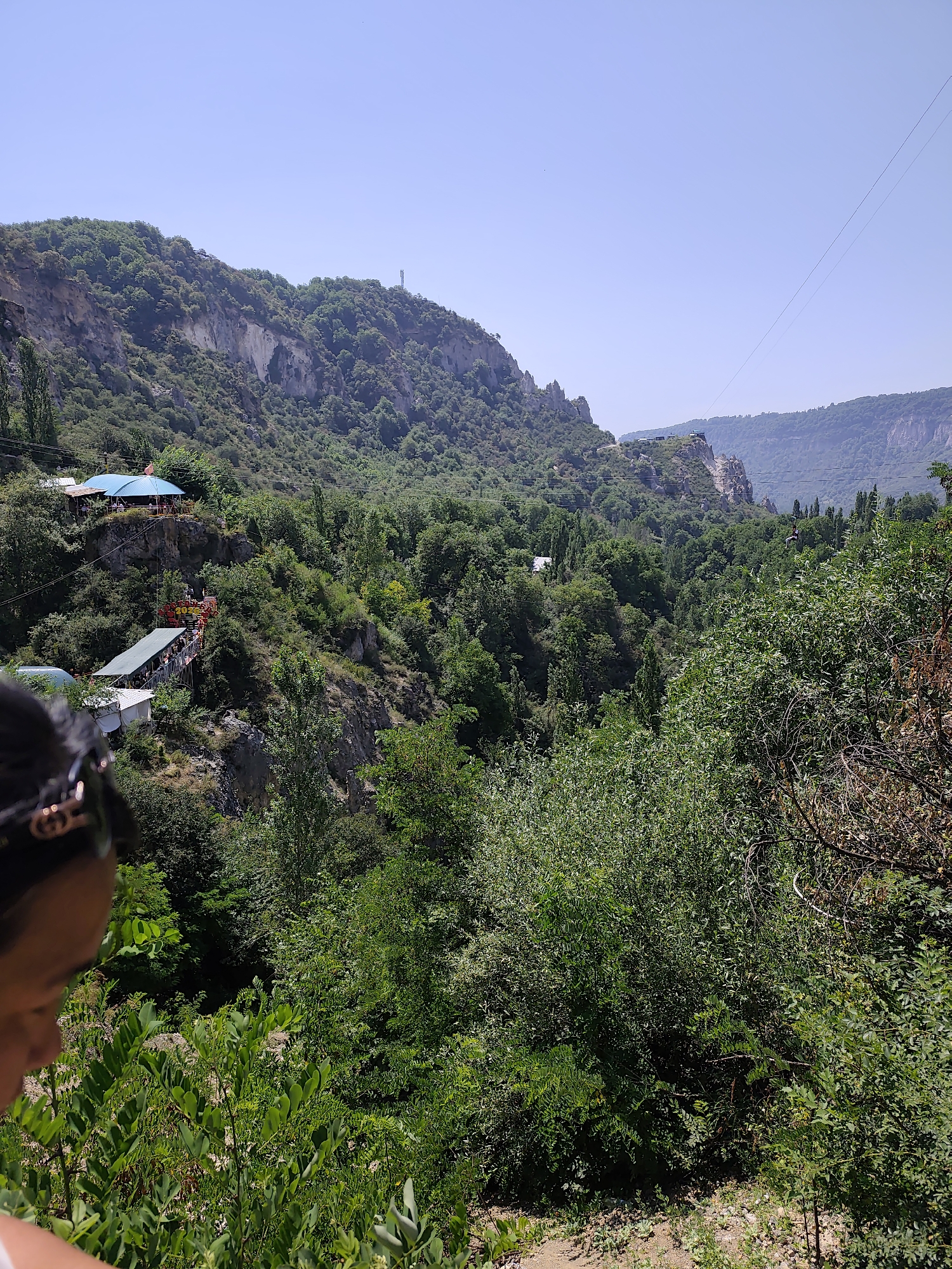The image captures a panoramic view of a mountainous landscape in Arstanbap, Kyrgyzstan, dominated by lush green trees covering the slopes. In the mid-ground, on a rocky outcrop, several structures with blue and green roofs are visible, suggesting a recreational area or small buildings. A prominent zipline stretches across the frame, with one gondola suspended mid-air. The foreground is partially occupied by the shoulder and back of a person wearing sunglasses, looking down towards the scenic vista. The sky is clear and blue, indicating a sunny day. The overall scene depicts a natural environment with tourist amenities, possibly an adventure park or a viewpoint.