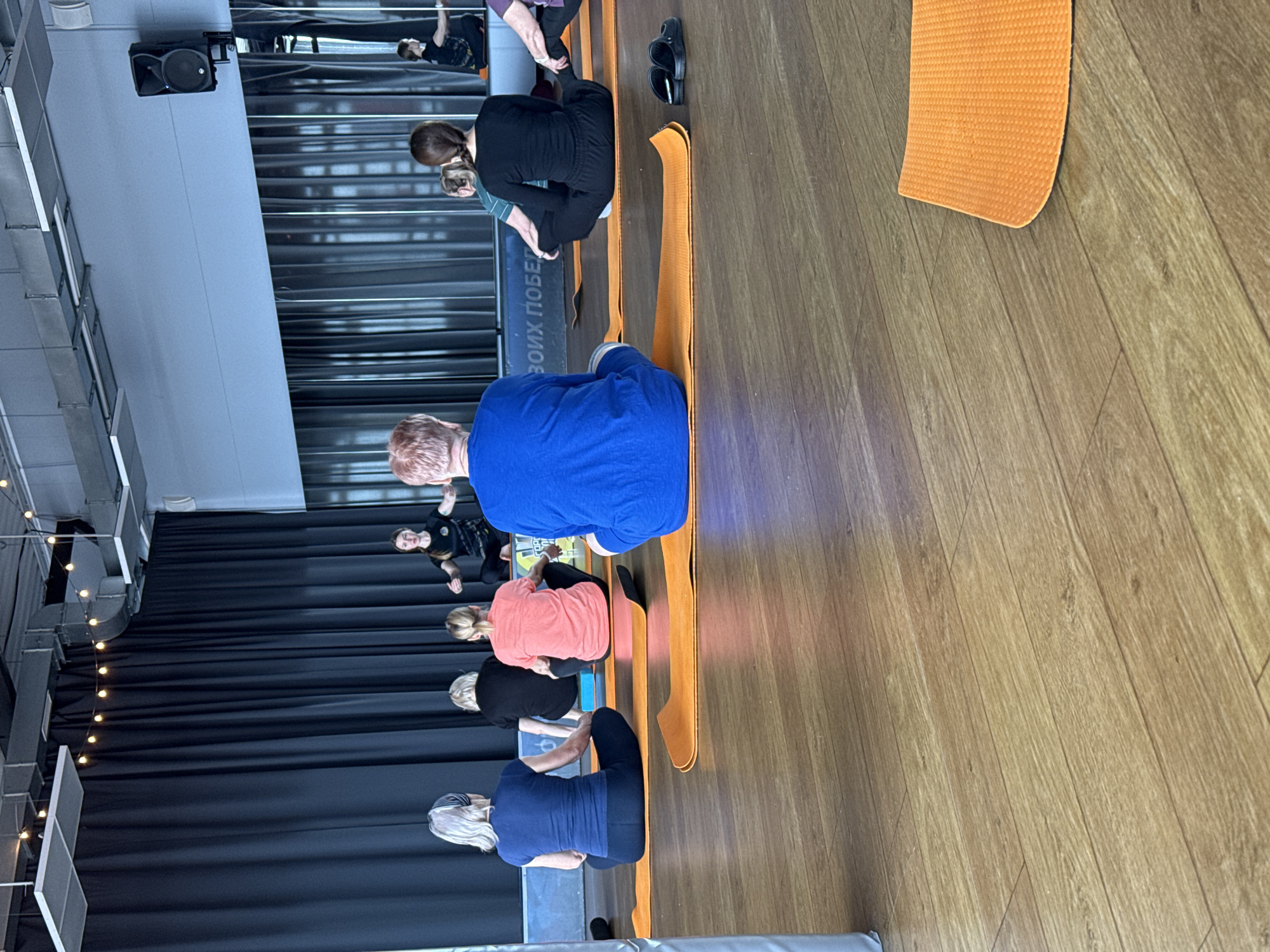 A group of at least seven people are sitting on orange yoga mats in a room. The location appears to be a studio or gym, with a black curtain in the background and a wooden floor. Some individuals are engaged in seated yoga poses. The room also contains a mirror showing a partial reflection of people, a speaker, and a wall with windows. The presence of text in Cyrillic on the windows indicates the location is in a Russian-speaking area. While the exact location in Votkinsk cannot be determined from this image alone, it is inferred to be in this city based on information available in the surrounding environment.