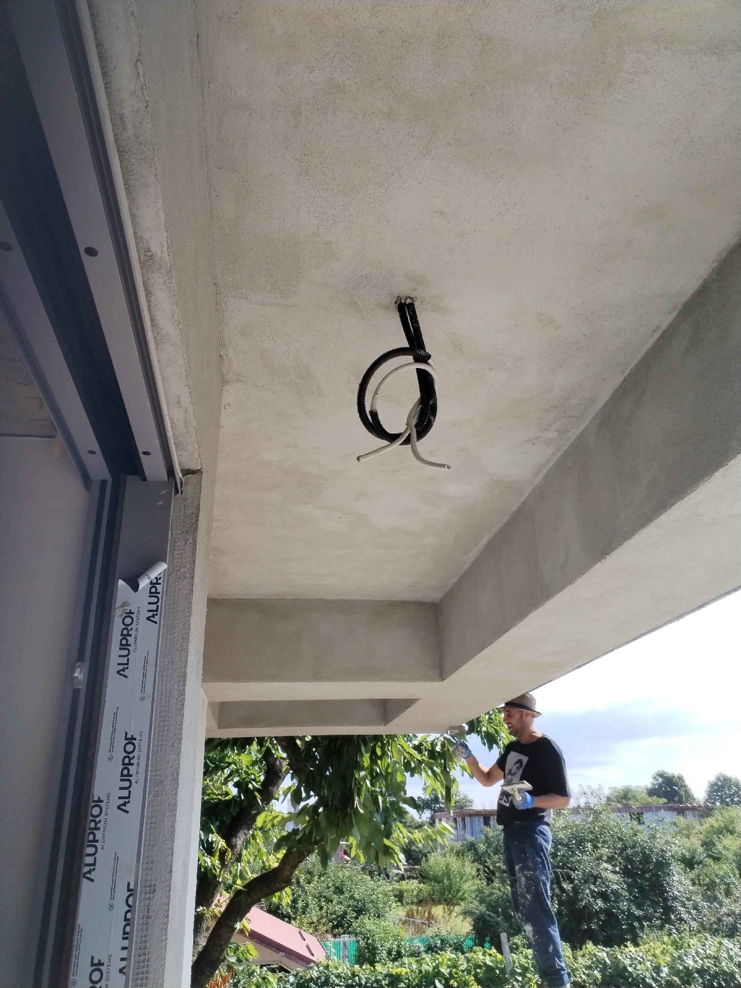 The image depicts an outdoor construction scene in Dumbrăveni, Romania. A man wearing a hat, a black t-shirt, blue jeans, and blue gloves is standing on a ladder, applying something with a brush to the underside of a concrete overhang. The man is holding another brush in his left hand. To the left of the man, a section of a building's facade is visible, with a window frame and some protective wrapping bearing the "ALUPROF ALUMINUM SYSTEMS" logo. Above and behind the man, the sky is visible with some clouds, and lush green trees and foliage form the background, suggesting a residential area. Electrical conduit and wires are visible suspended from the concrete ceiling of the overhang. The overall impression is a snapshot of exterior renovation or construction work.