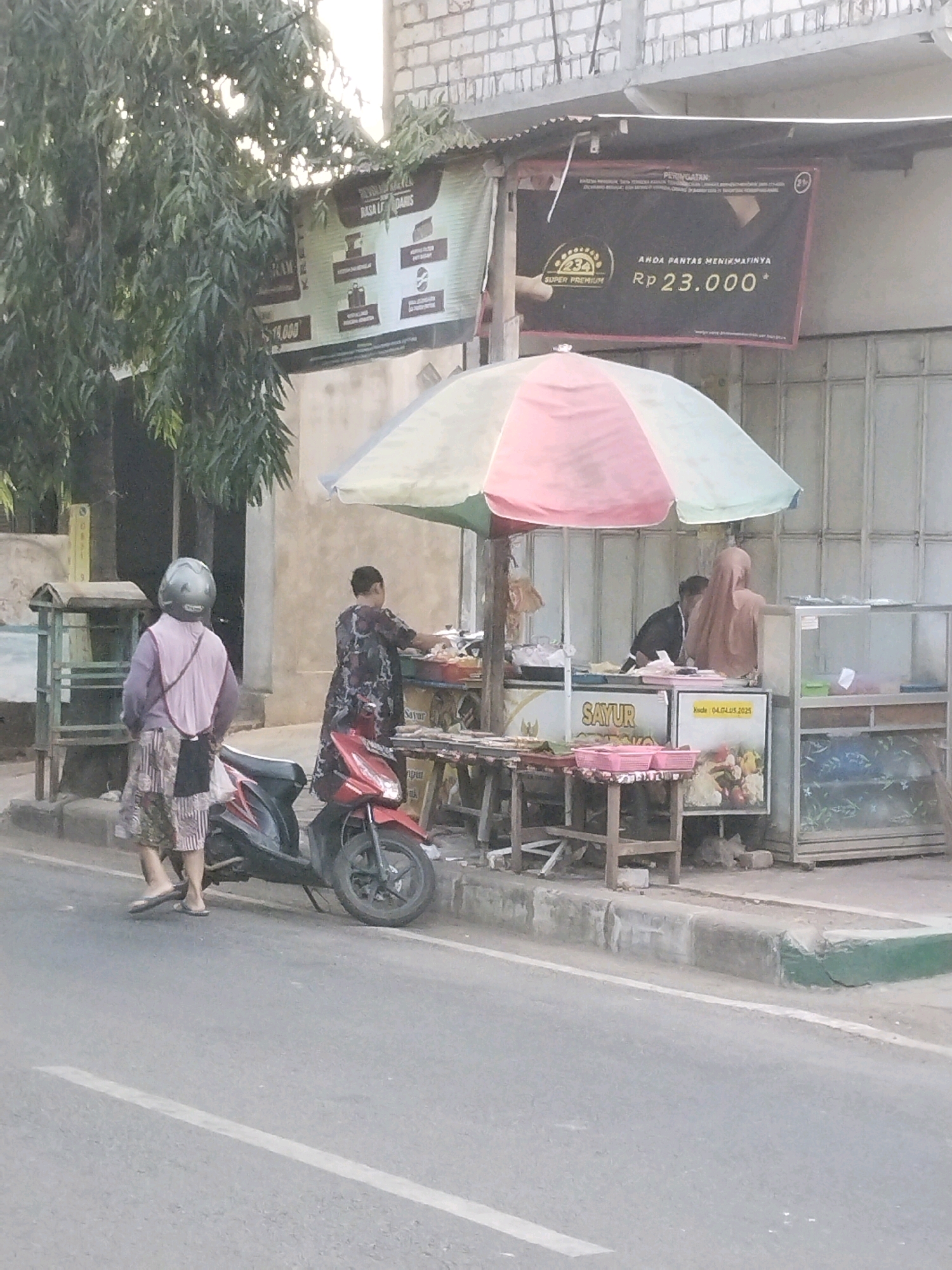 This image depicts a street scene in Semanding, Indonesia, near a food stall. In the foreground, a red motorcycle is parked on the side of the road. A person wearing a pink shirt, a patterned skirt, and a helmet stands near the motorcycle, facing away from the camera. Behind the motorcycle, a food stall is set up with an umbrella providing shade. A woman in a dark patterned top is working at the stall, preparing food. Another person, wearing a light brown hijab, is also visible at the stall. The stall is decorated with banners and displays of food items, including vegetables and what appear to be packaged goods. A glass display case is visible on the right side of the stall. In the background, a building with a tiled roof and brick walls is present. A large green leafy tree is to the left of the scene. A black banner with text and logos is hanging above the stall. The environment suggests a commercial area with food vendors.