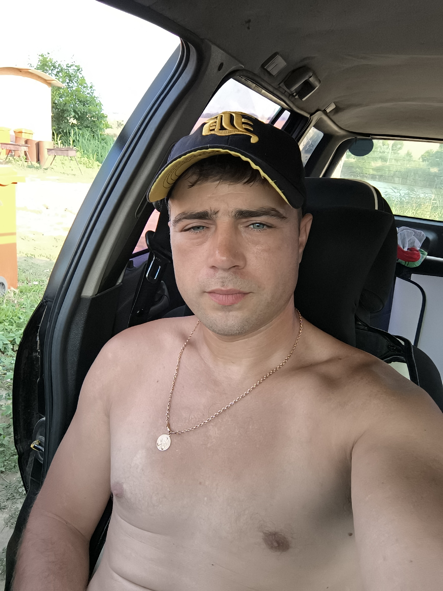 A shirtless man wearing a black and yellow baseball cap sits in the driver's seat of a car. He has a gold chain with a round pendant around his neck. The car appears to be parked outdoors, with greenery and a small building visible in the background. The lighting suggests it is daytime.