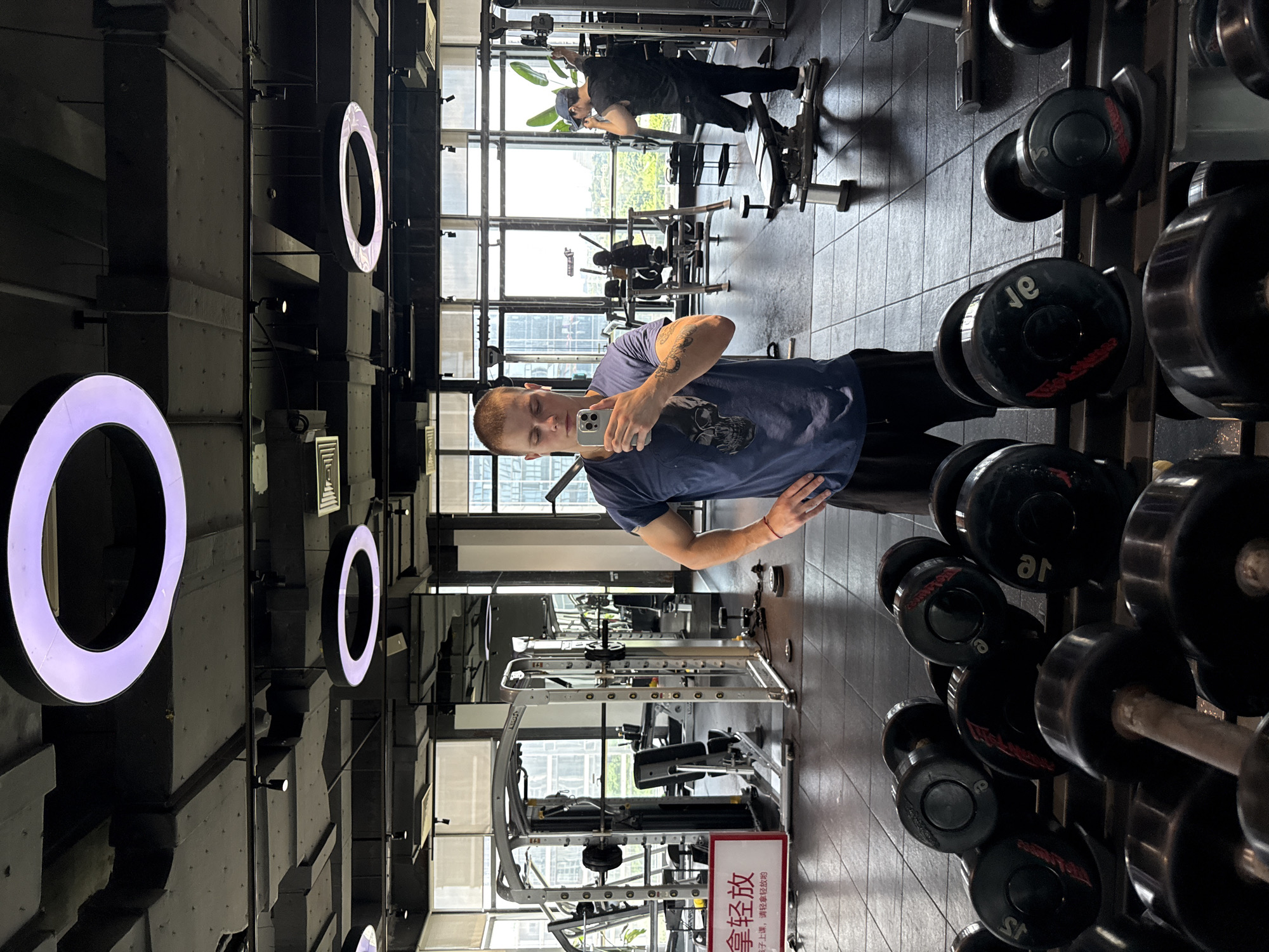 A young man with short blonde hair and a skull graphic t-shirt takes a mirror selfie in a gym. He is standing in front of a rack of dumbbells. In the background, another man in a black t-shirt and blue cap is working out on a weight machine. The gym has large windows with a view of a city skyline, suggesting the location is Haizhu District, China. There are other gym equipment and circular light fixtures visible in the ceiling.
