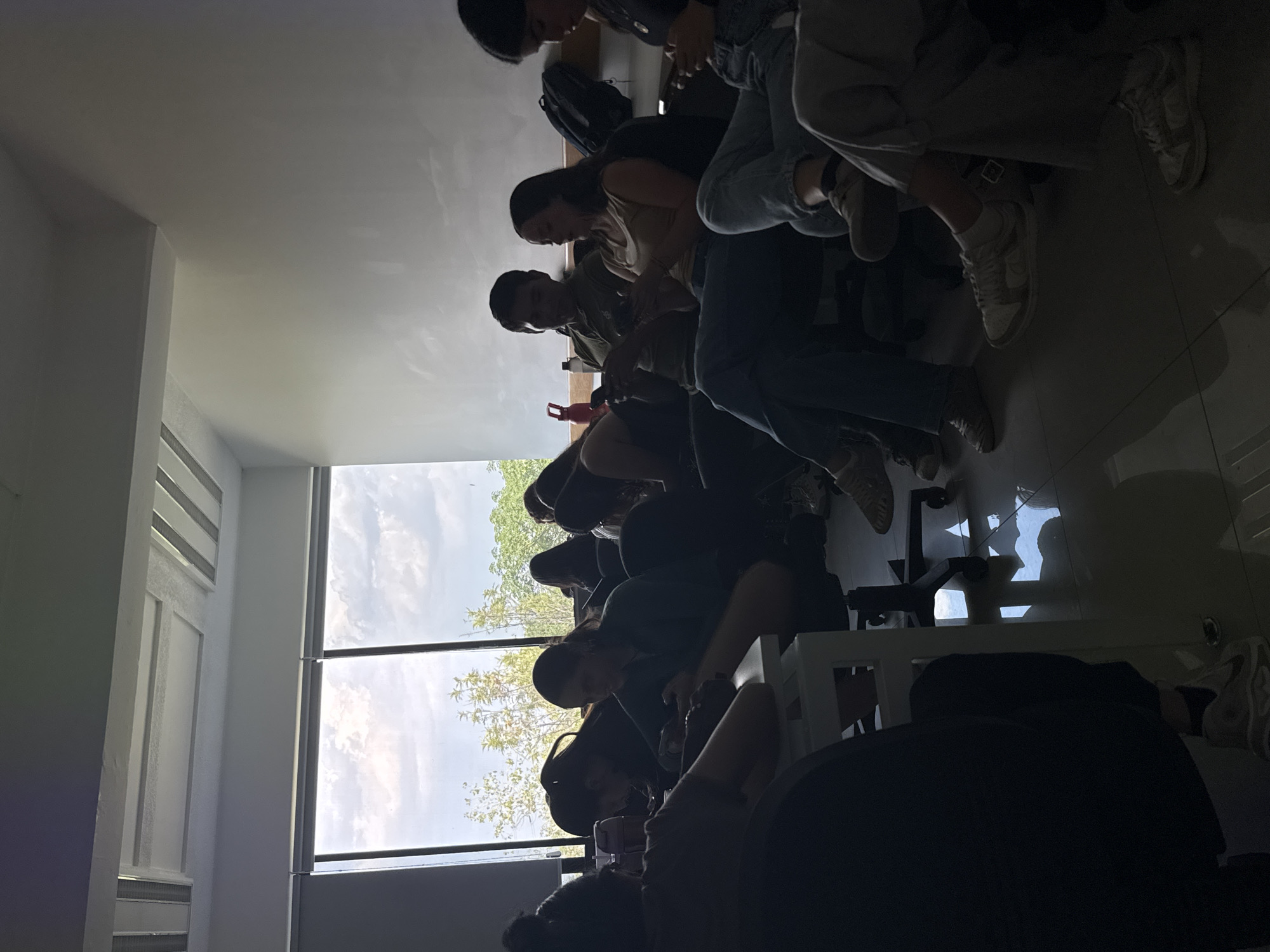 The image depicts a group of people sitting indoors, possibly in a classroom or waiting area. They are mostly looking down, seemingly occupied with their phones. A large window provides natural light and a view of trees and sky outside. The interior has a modern design with white walls and tiled floors. The location is San Andrés Cholula, Mexico.