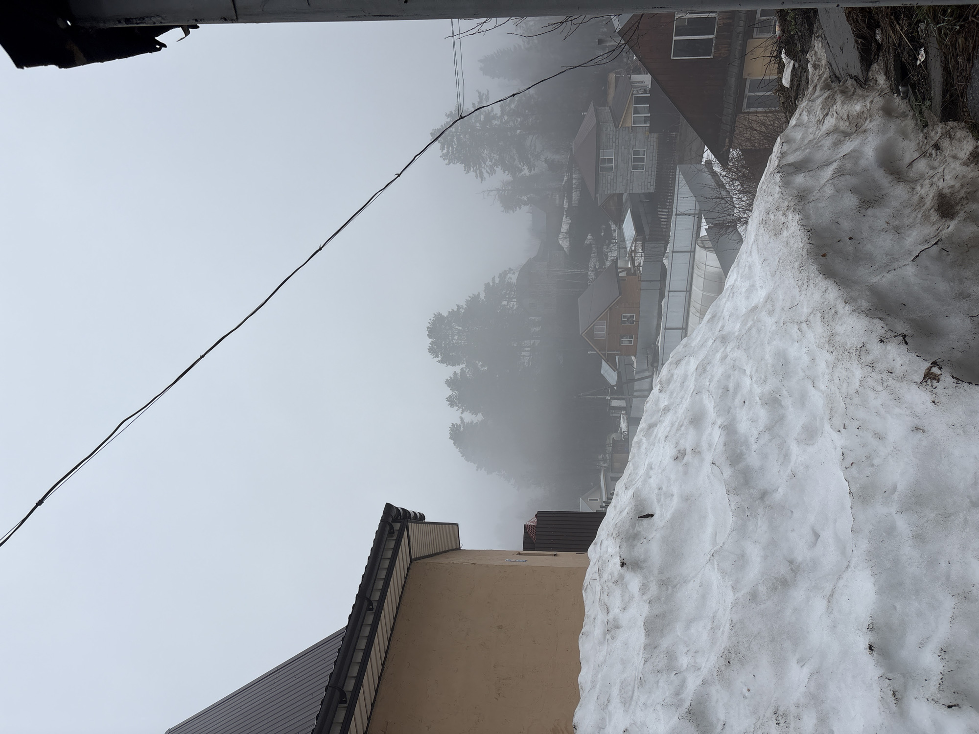 A daytime, medium-shot, medium-angle photograph depicts a hazy, foggy scene in Chaikovsky, Russia. Visible are buildings and trees shrouded in mist, as well as a significant accumulation of snow on what appears to be a rooftop or ledge. A utility line stretches across the upper left portion of the frame. The architectural features suggest residential buildings. The overall atmosphere is muted due to the heavy fog.