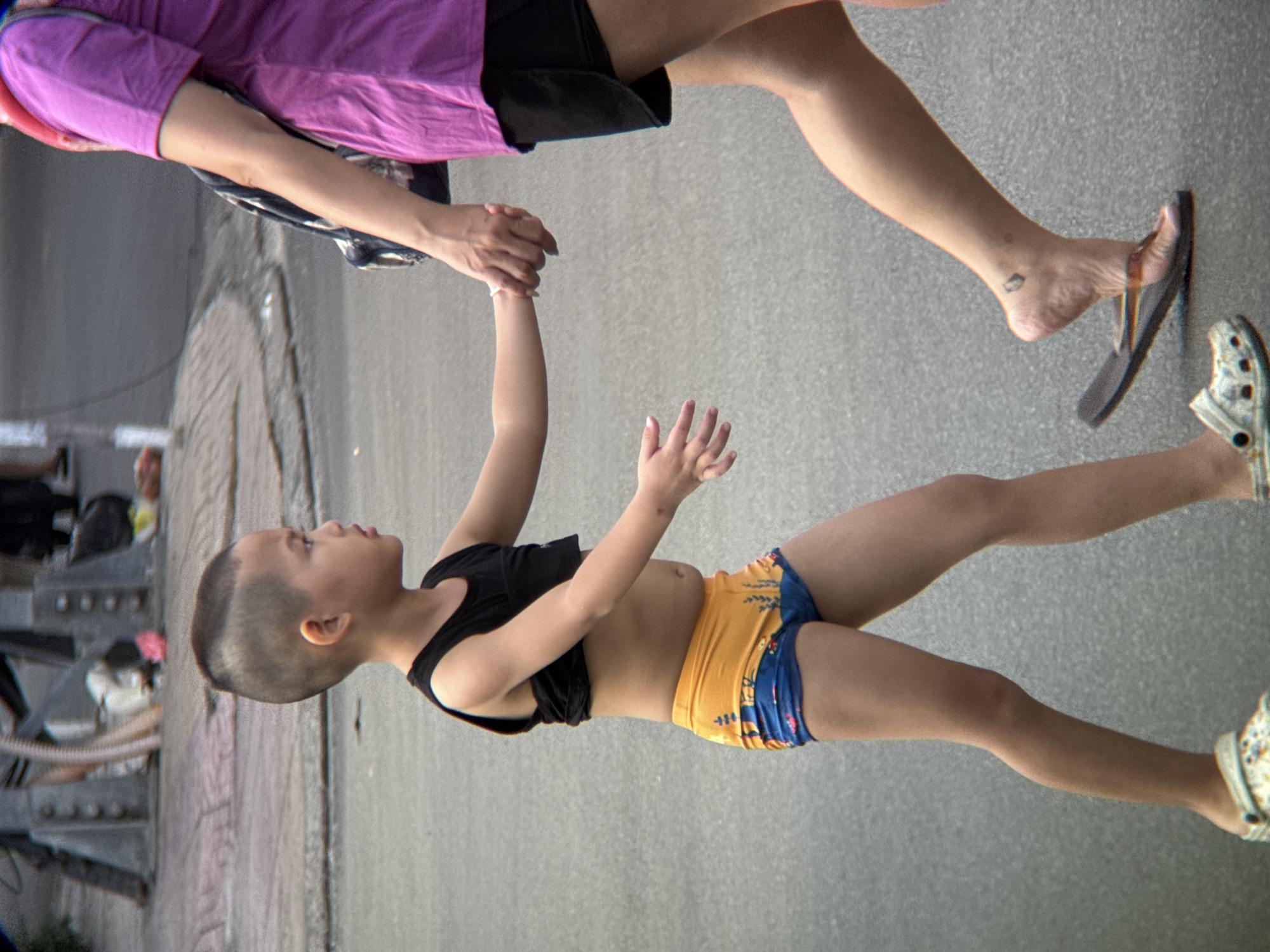 A young boy and an adult, likely his mother, are walking on a paved street in Nha Trang, Vietnam. The boy, facing left, wears a black tank top and orange and blue patterned shorts. He is holding the adult's hand with his left hand. The adult, mostly visible from the waist down, wears a purple t-shirt and black shorts, and carries a backpack. Her right hand is holding the boy's hand. Both are wearing open-toed footwear. The background shows a sidewalk with some debris and a blurred structure that appears to be part of a building or street furniture. The ground is asphalt.