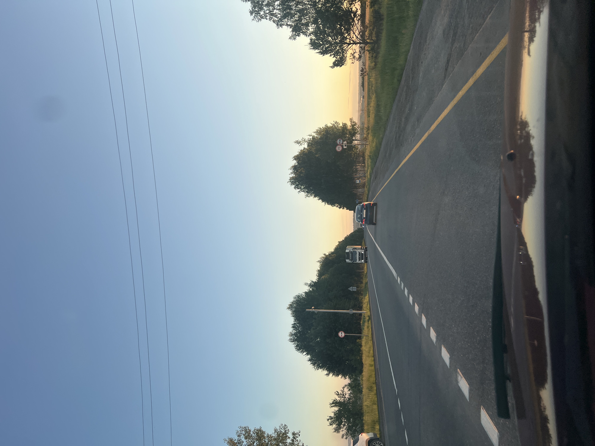 The image depicts a tranquil view from inside a vehicle traveling along a rural highway at either dawn or dusk, indicated by the warm, golden light on the horizon transitioning to a cool blue sky overhead. The scene shows an asphalt road extending into the distance, with a solid yellow line marking the left edge and a dashed white line separating the two lanes further ahead.
Several subjects are visible on the road: a dark-colored car and a white semi-truck are driving away from the camera in the right lane. On the far right, a dark, reflective surface of the vehicle from which the photo was taken is prominent. On the far left, a portion of a white car's wheel and body can be seen, possibly parked off the shoulder.
The road is flanked by dense foliage and trees on both sides, with patches of green grass visible beyond them. In the distance, a hazy landscape stretches towards the horizon. Power lines cut across the upper left section of the sky.
Notable details include two road signs on a pole to the right of the road: a red circular sign with a diagonal white line, often indicating a prohibition (such as "no entry" for certain vehicles), and below it, a rectangular white sign with a red border displaying the number "70," signifying a 70 km/h speed limit. A faint, dark circular smudge is visible in the upper left sky, likely a camera lens or sensor dust spot. The overall impression is that of a peaceful journey through the countryside near Semenovo, Russia, during a scenic time of day.