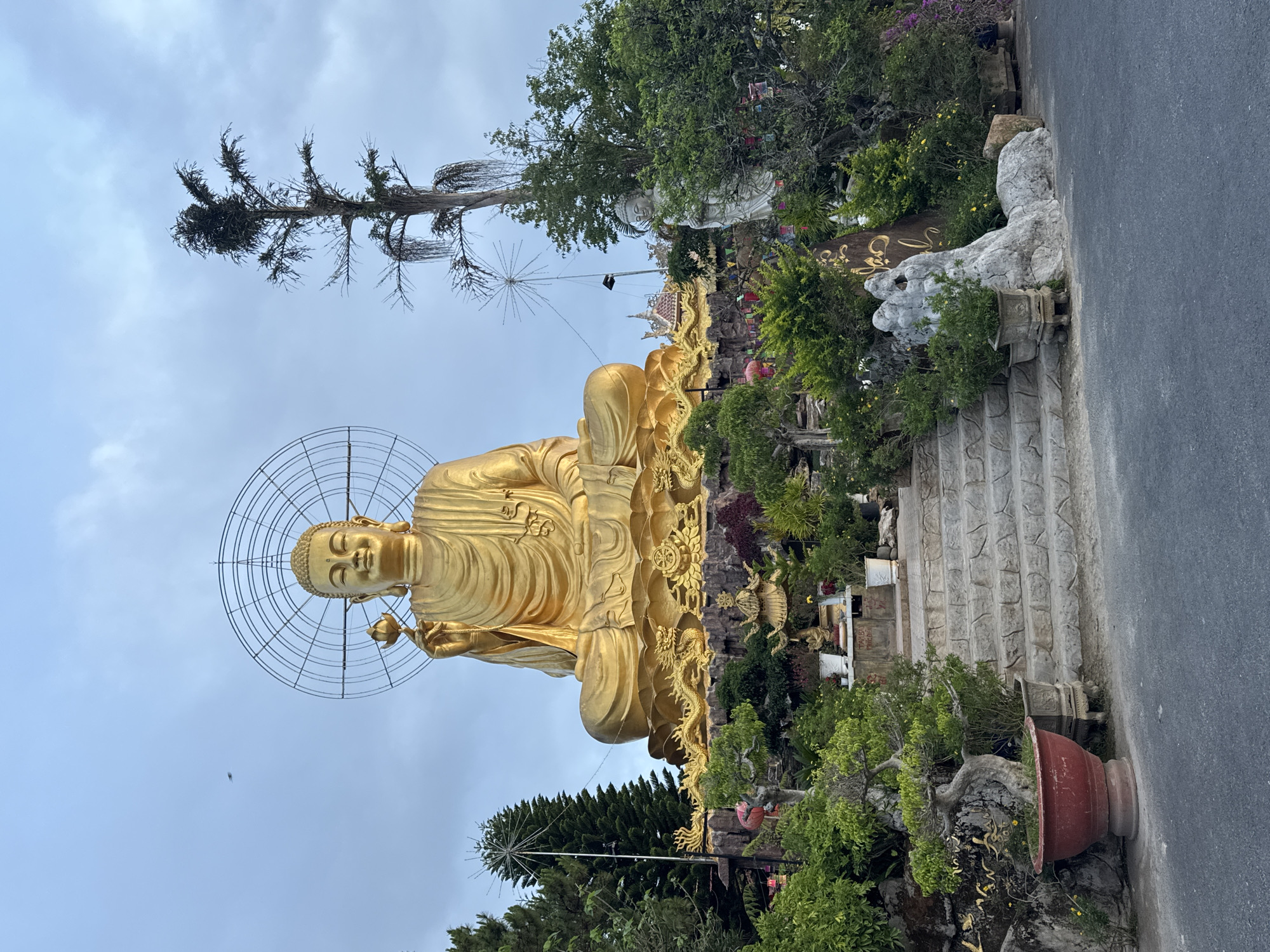The image shows a large, golden Buddha statue seated in the lotus position. It is located in Lâm Viên, Đà Lạt, Vietnam. The statue has a circular halo-like structure behind its head. The statue is surrounded by greenery, trees, and some architectural elements. There are stairs leading up to the statue. Part of a paved road is visible on the right side. The sky is overcast. Another smaller white statue can be seen above the golden one, as well as various signs and decorations.