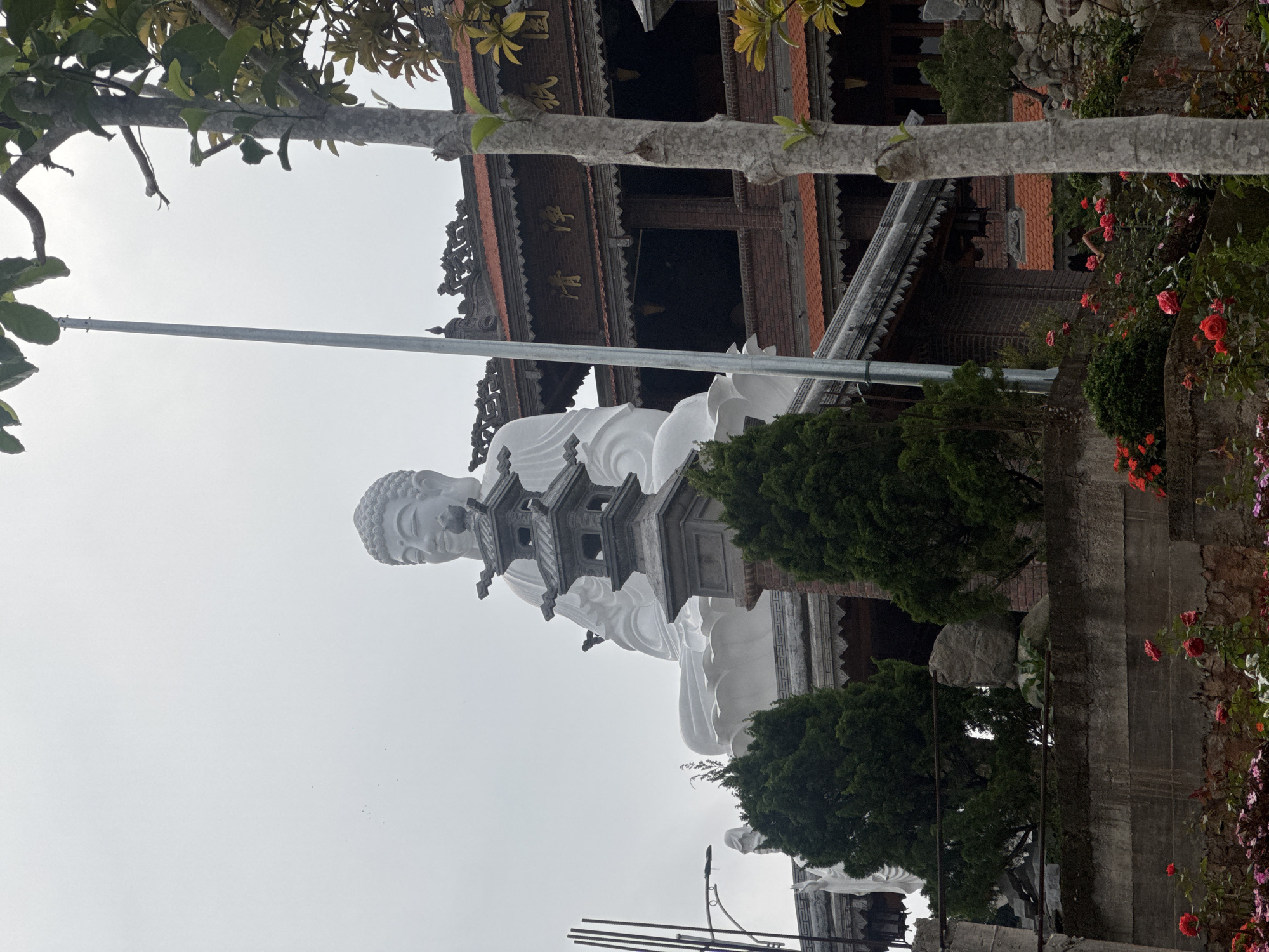 A monumental white statue of Buddha is the focal point, flanked by trees and a pagoda-style structure. The location is Da Lat, Vietnam, recognized by the architectural style. The statue is intricately detailed, from the curls of the hair to the folds of the robe. In front of the Buddha statue is a gray pagoda statue. It is surrounded by greenery, including manicured bushes and trees. Red and purple flowers and plants can be seen on the right side of the frame, growing along a set of stone steps. The pagoda-style building, with its multi-tiered roof and decorative carvings, provides a backdrop, adding to the religious atmosphere. A gray pole extends from the bottom center and goes over the statue to the top left. The sky is overcast, creating a soft, diffused light that bathes the scene.