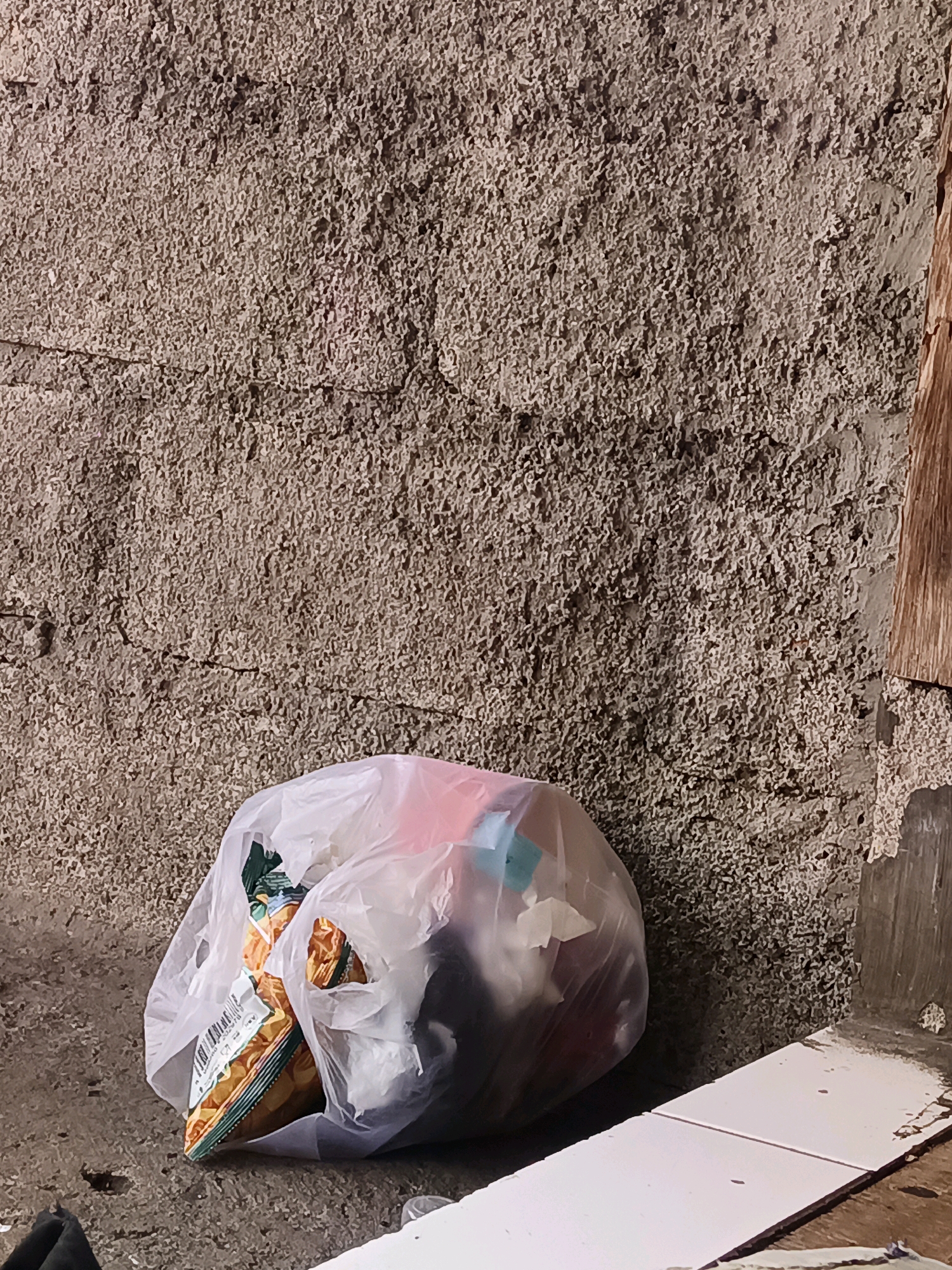 In the city of South Jakarta, Indonesia, a plastic bag filled with various items, including what appears to be snacks and other colorful objects, rests on a concrete floor against a textured, rough-hewn wall. The wall exhibits a coarse, granular surface with visible cracks, suggesting age and potential disrepair. To the right of the wall, a portion of a vertical wooden plank is visible, also showing signs of wear. In the foreground, two white rectangular tiles are laid out on a wooden surface, with a small amount of dirt or debris scattered on the floor around them. The lighting in the scene appears to be natural, casting shadows that highlight the textures of the wall and the contents of the plastic bag.