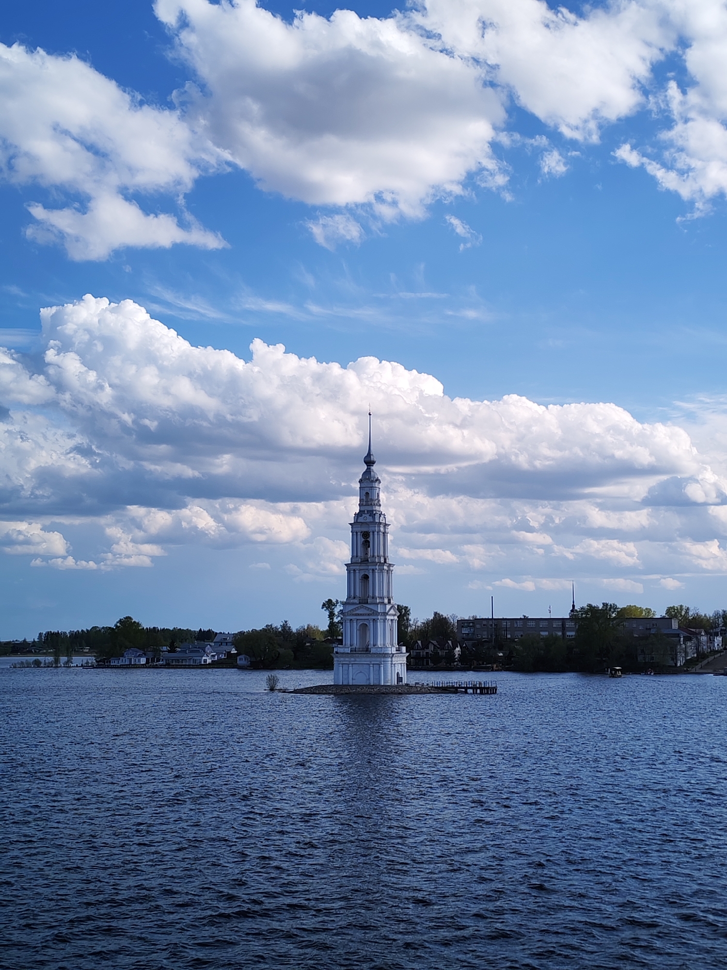The image shows a solitary white bell tower standing in the middle of a body of water, likely a lake or reservoir, in Kalyazin, Russia. The tower, seemingly isolated and surrounded by water, rises prominently against a backdrop of a partly cloudy blue sky. In the distance, the shoreline with some trees, houses, and buildings are visible on both sides of the tower. The water surface has ripples and waves. The sky is filled with fluffy white clouds and patches of clear blue.