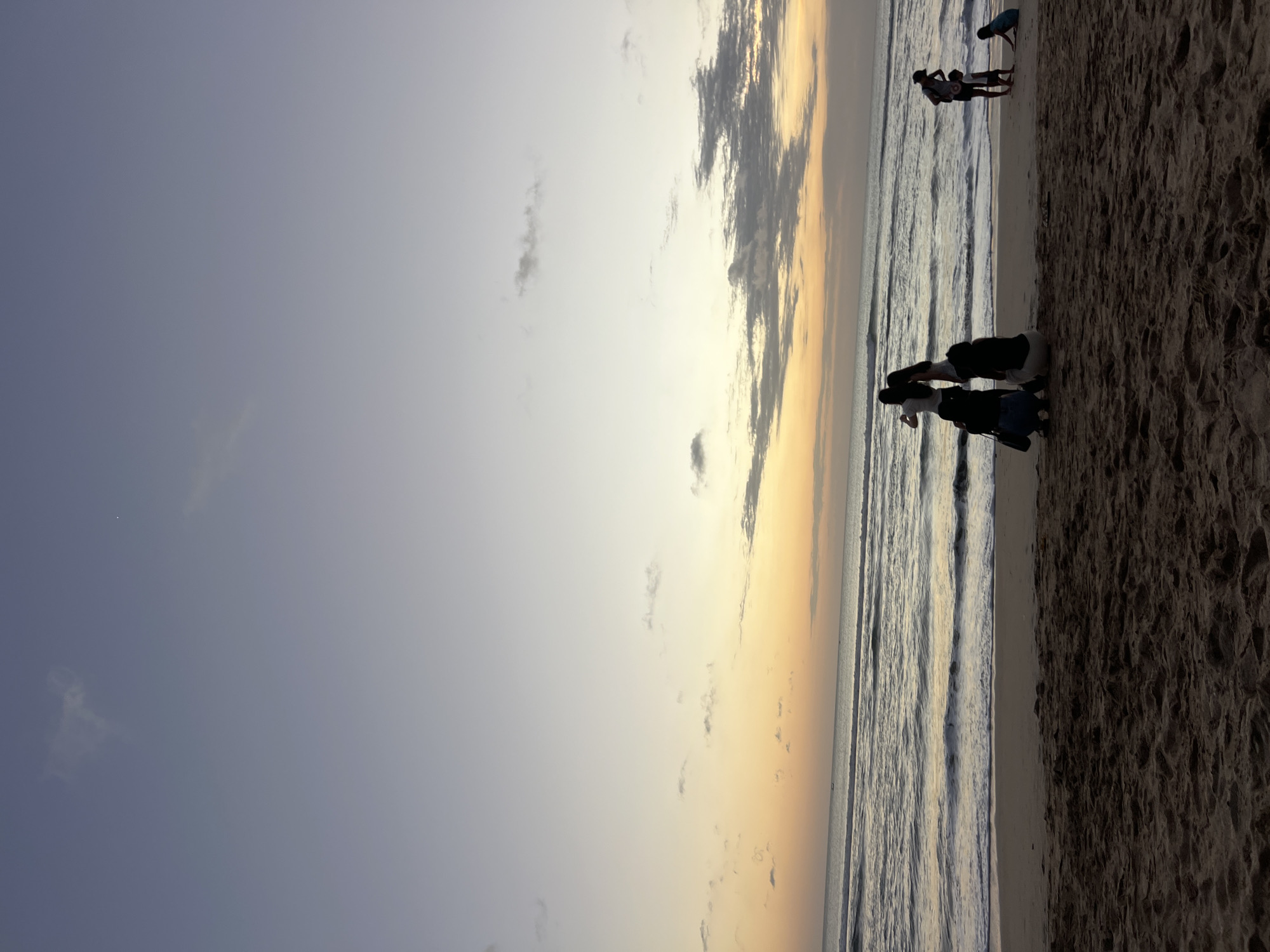 The image depicts a beach scene in Kuta, Indonesia, at dusk. The sky displays a gradient of soft purples and oranges, with scattered clouds. Gentle waves are lapping onto a sandy shore. In the foreground, the sand is textured and uneven.
On the beach, several individuals are present. To the left, a group of four people are gathered near the water's edge. Two individuals are standing and appear to be taking a photo or looking at a device, while two others are crouching. To the right, a person in a hat stands with a young child, and further to the right, a child is seated on the sand. The overall atmosphere suggests a peaceful evening on the coast.