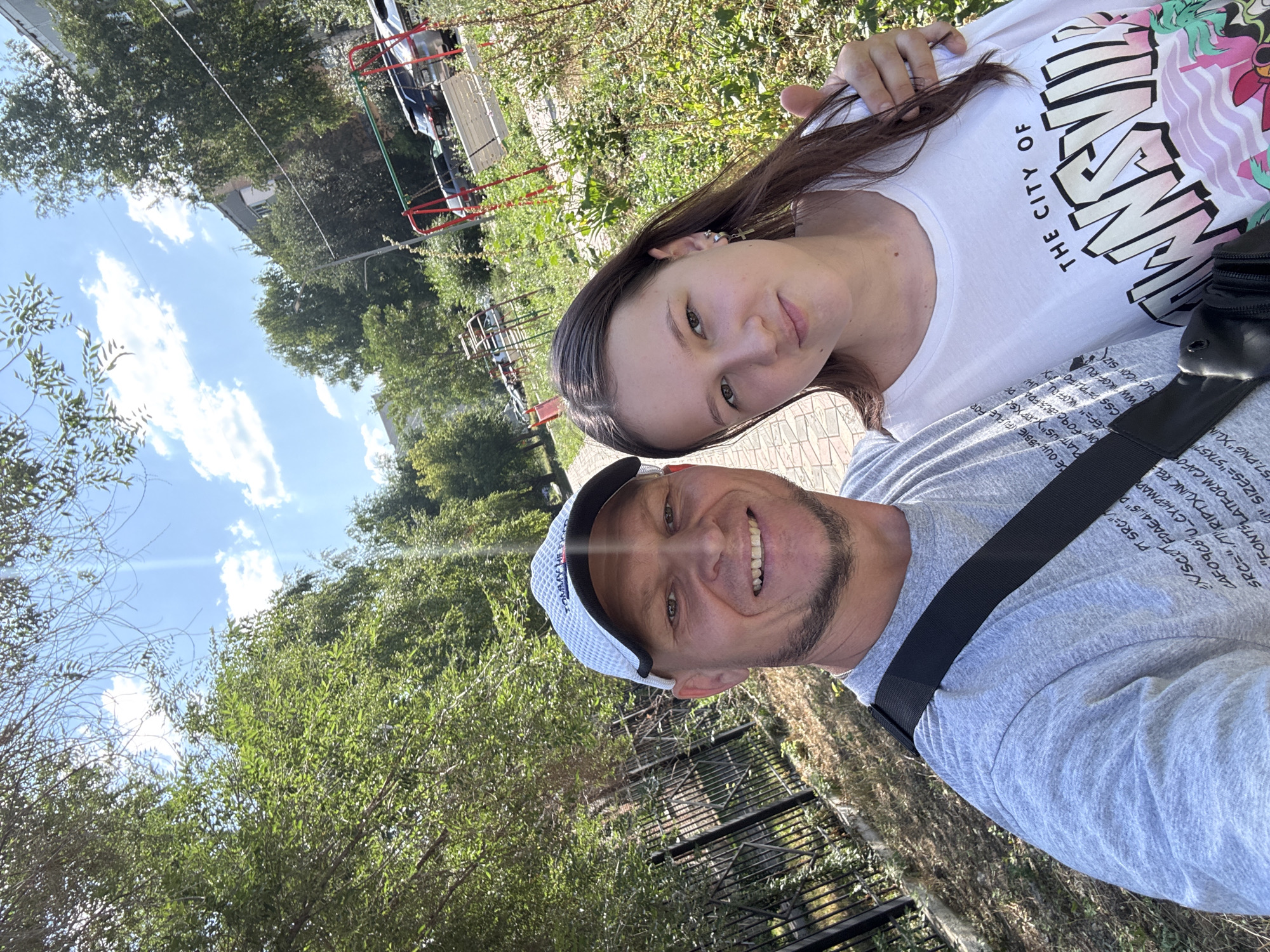 In a sunlit outdoor scene, a man and a young woman are captured in a selfie. The man, wearing a light-colored cap and a gray t-shirt, smiles broadly at the camera. The woman, with long brown hair, is dressed in a white t-shirt and places her right arm around the man's shoulders, giving a slight thumb-up gesture. They are positioned on a paved walkway, with a black metal fence to their left and lush green trees and vegetation to their right. In the background, a playground structure with red frames is visible, along with parked cars and a multi-story building. This scene appears to be a residential courtyard or park area within the city of Oskemen, Kazakhstan.