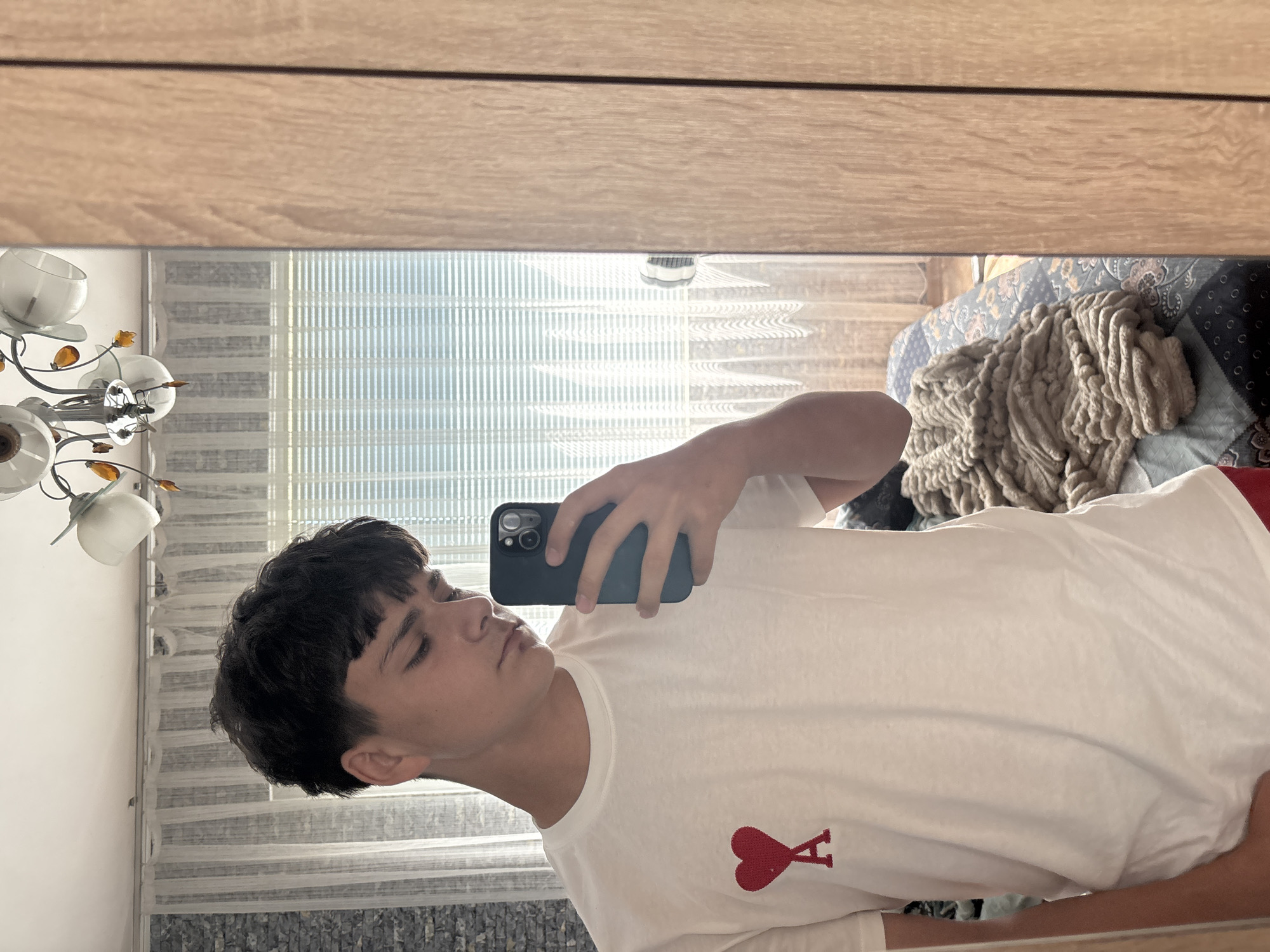A young man is taking a selfie in a mirror indoors. He is wearing a white t-shirt with a red heart logo on the chest. He is holding a dark-colored smartphone in his left hand. The background shows a room with blinds on the window, a chandelier, and a bed with blankets. The location in the city of Kardzhali, Bulgaria is unknown and cannot be determined from the image.