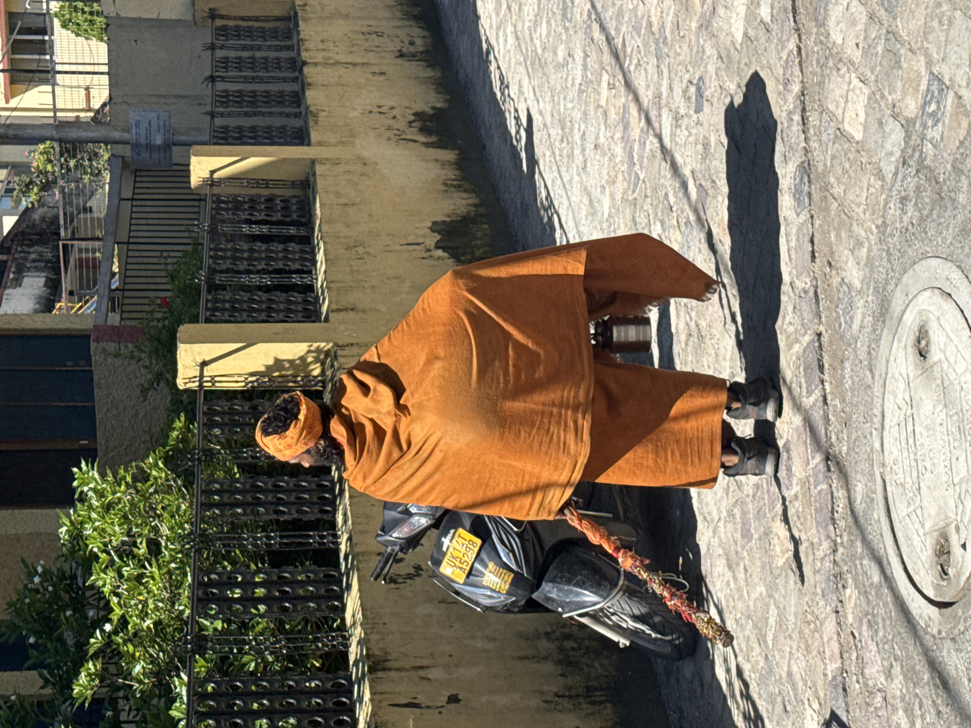 A man dressed in an orange robe and turban stands on a cobblestone street in Rishikesh, India. He is facing away from the camera and holding a decorated staff. A black scooter is parked to his left. The street is lined with a yellow wall and a black metal fence. Sunlight casts shadows on the ground.