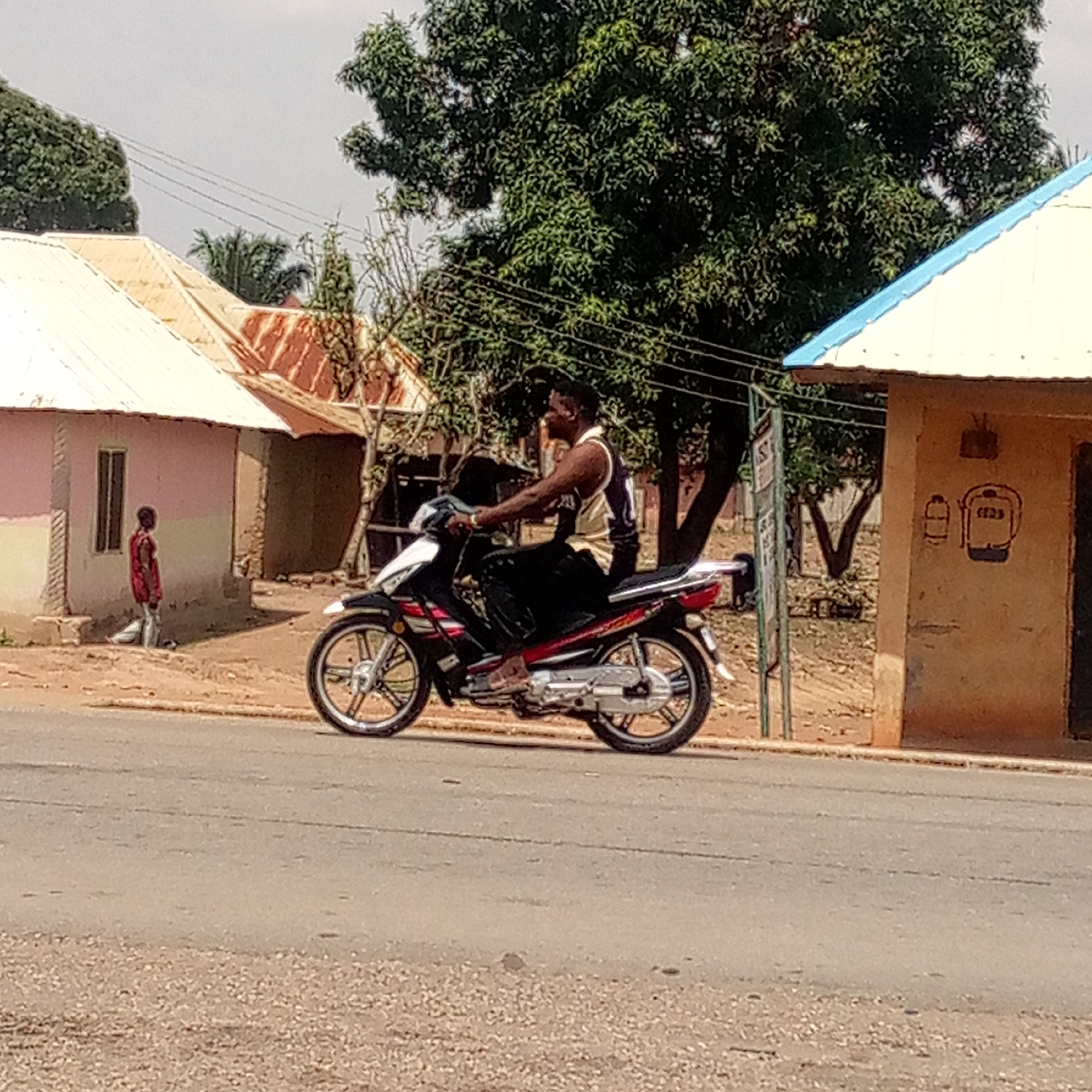 A man rides a black and red motorcycle on an asphalt road in Unknown, Unknown. The location appears to be a rural area or small town, indicated by the presence of modest buildings with corrugated metal roofs and dirt yards. The man is wearing a sleeveless shirt, and dark pants, and is not wearing a helmet. The background includes a child standing near a pink-colored building and trees that appear to be providing shade. On the right, there is a small structure with a blue-edged roof, seemingly a shed or a bus stop, and signs indicating the structure is a bus stop. Power lines can be seen strung overhead.