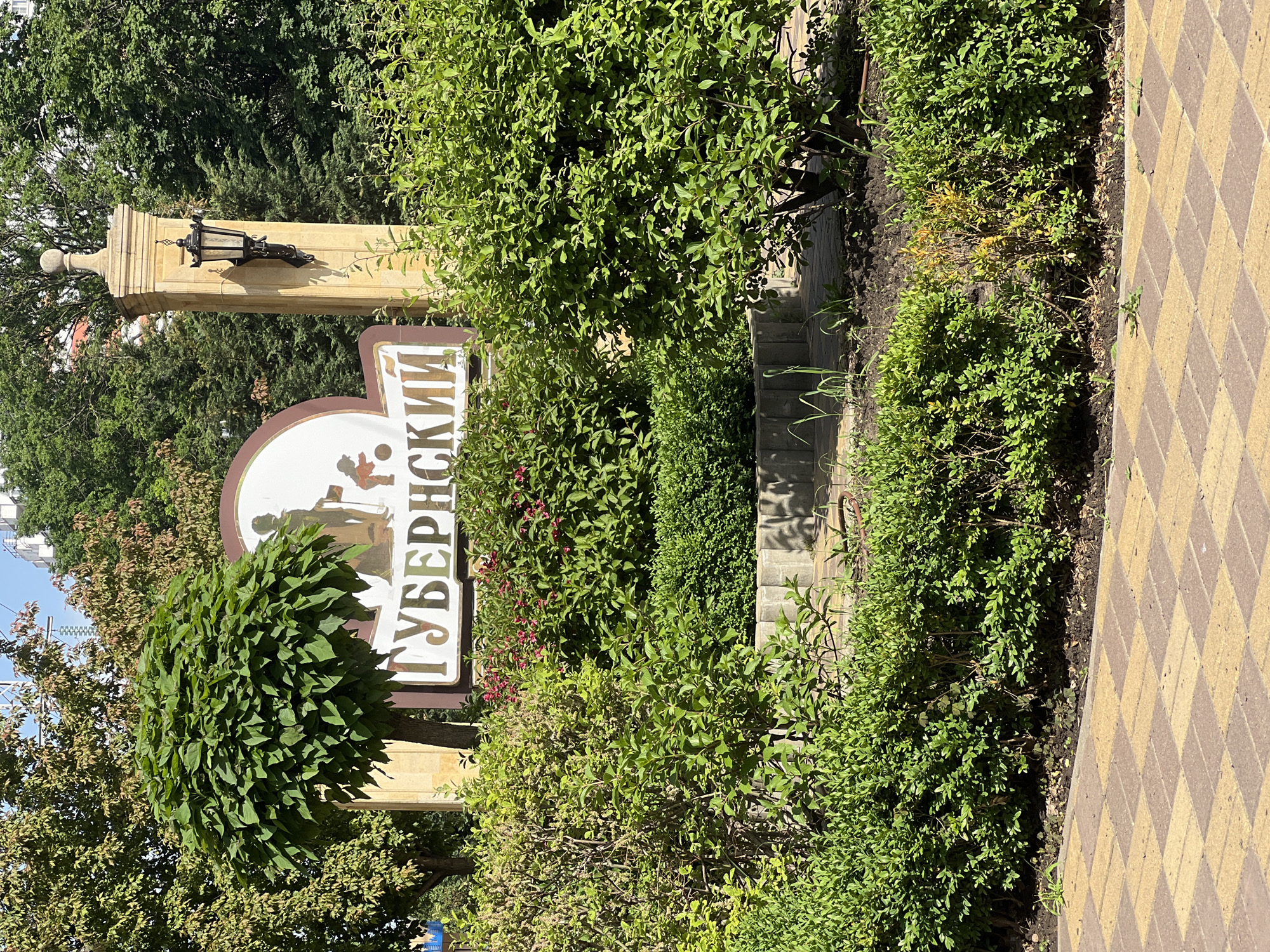 The image shows an outdoor scene in the city of Krasnodar, Russia, featuring a sign that reads "Губернский" ("Gubernskiy"). The sign is partially obscured by dense greenery and bushes. The sign itself is mounted on a structure that includes a lamp post. A paved walkway with a diamond pattern design runs alongside the greenery. There are steps leading up into the foliage. The scene is set during the day with good lighting.