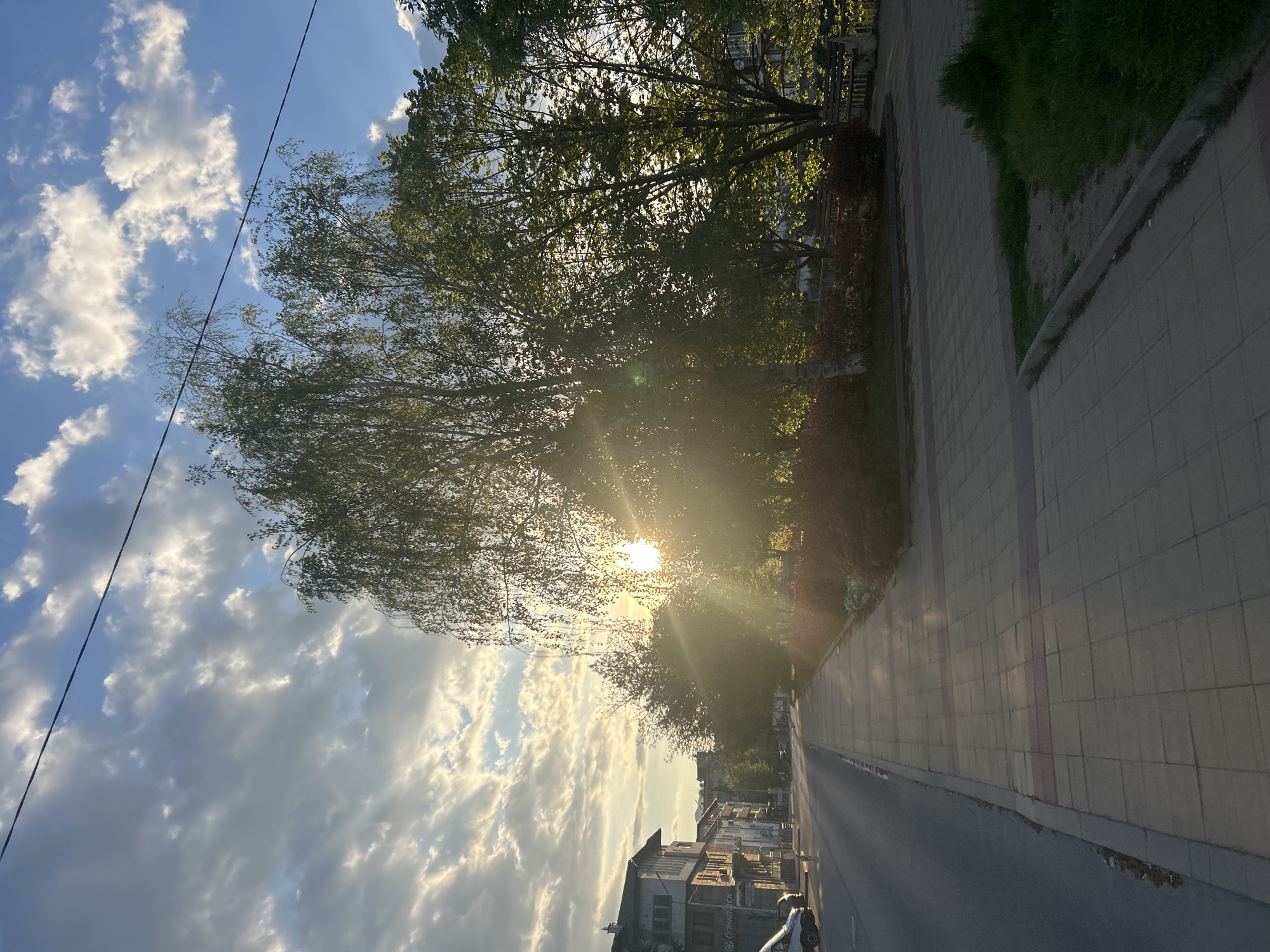 The image depicts a sunlit street scene in Zlatograd, Bulgaria. The perspective is from a slightly elevated position, looking down the street. The road stretches into the distance, flanked by buildings on the right side, the closest of which is multi-storied, of a light color. Along the right of the road is a pedestrian walkway. Large trees line the sidewalk. The sun is partially obscured by the trees, creating a lens flare effect. To the left is a view of the sky filled with clouds. A power line runs across the top left of the frame.