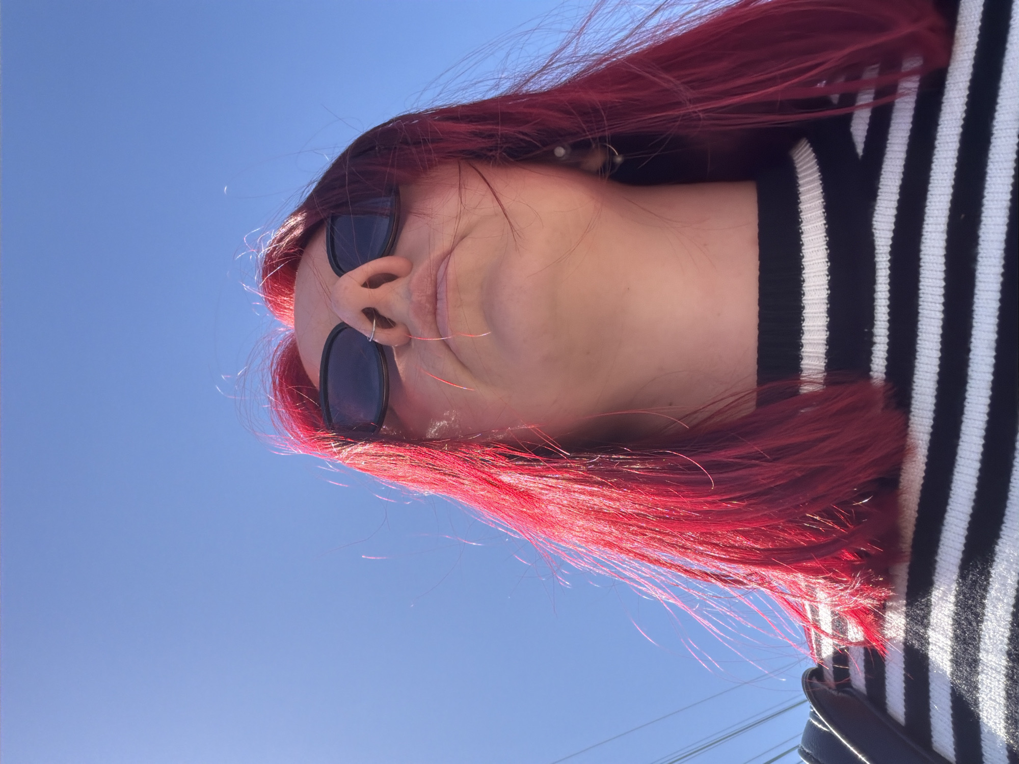 A low-angle, vertically-oriented, color photograph shows a person with red hair wearing black sunglasses and a black and white striped sweater. A silver nose ring is visible. A clear blue sky is in the background. The location is not discernible.