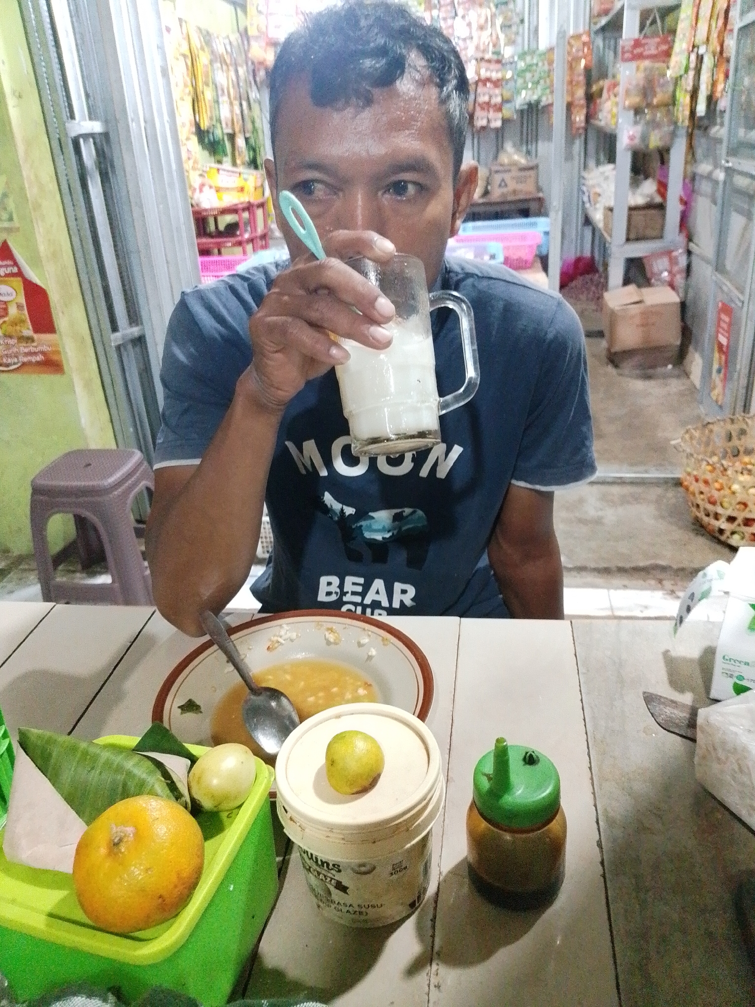 In Kecamatan Pati, Indonesia, a man is seated at a table, holding a glass of a white beverage and a spoon. The man is wearing a dark blue t-shirt with a graphic design. In front of him, on the table, is a plate with food and a spoon, a green plastic container with a round orange fruit, a wrapped item, and a small yellow fruit. To the right of the plate is a white container with a green cap and a small yellow fruit on top, and a small sauce dispenser with a green cap. The background shows shelves stocked with various items, suggesting the location is a shop or market stall.