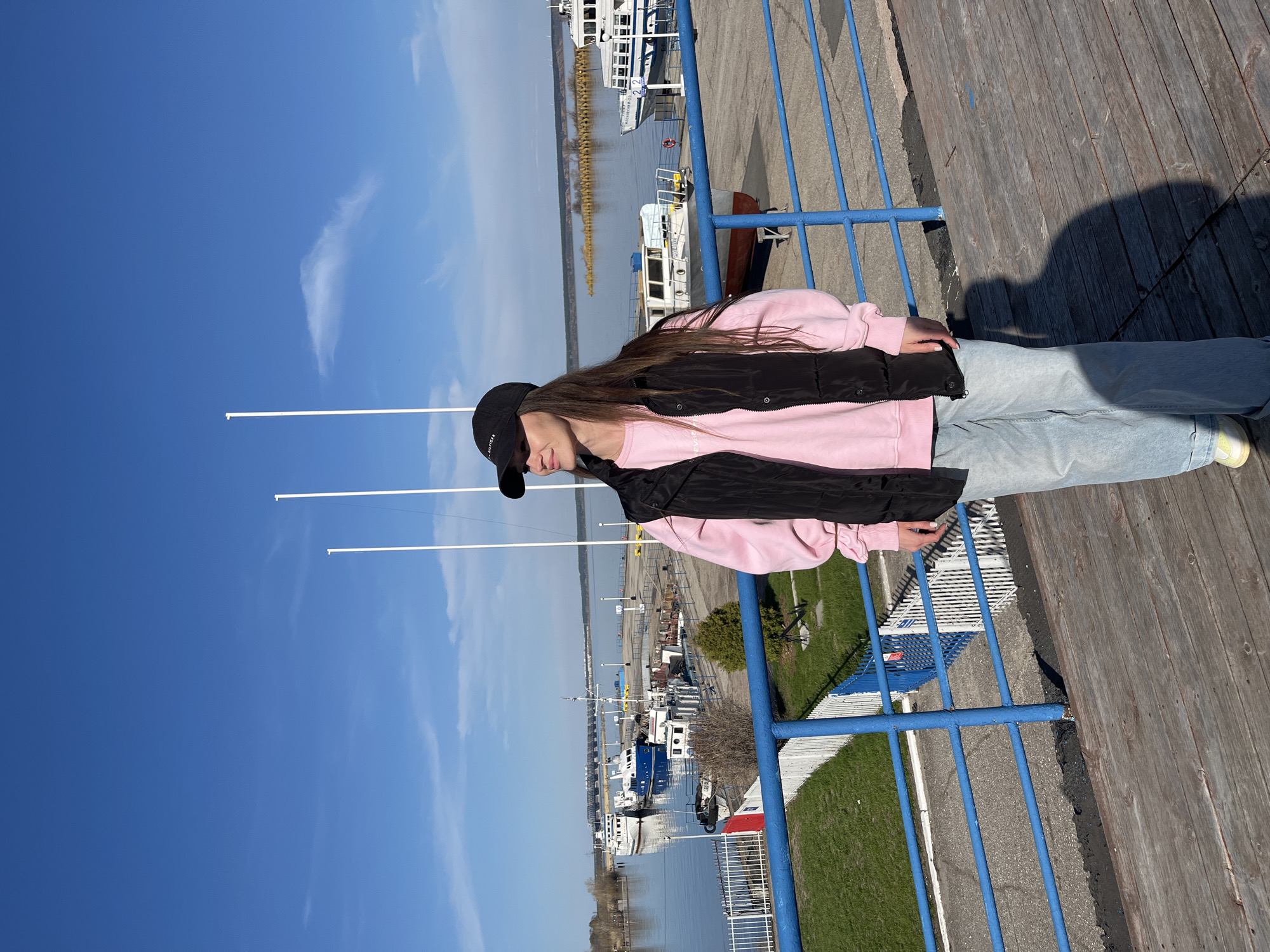 A young woman stands on a wooden deck overlooking a waterfront in Ulyanovsk, Russia. She is wearing a pink sweatshirt, black vest, light blue jeans, and a black baseball cap. The waterfront is visible with docked boats, grassy areas, and buildings in the distance. A blue railing runs along the edge of the wooden deck. The sky is blue with some light clouds.