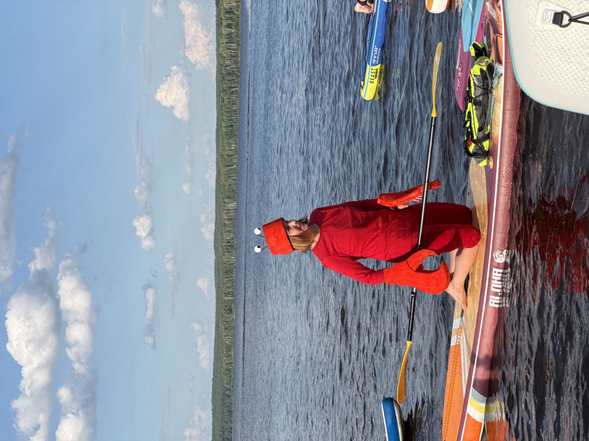In the city of Chaikovsky, Russia, on a lake with a tree line in the background, a woman dressed as a lobster kneels on a paddleboard. She wears a red outfit with large red lobster claws attached to her hands and a red hat with two googly eyes. She holds a paddle across her lap. Other paddleboards are visible on the water, one yellow and blue with the word "BEAST" printed on it, and another with a life vest on it. The sky is blue with white clouds.