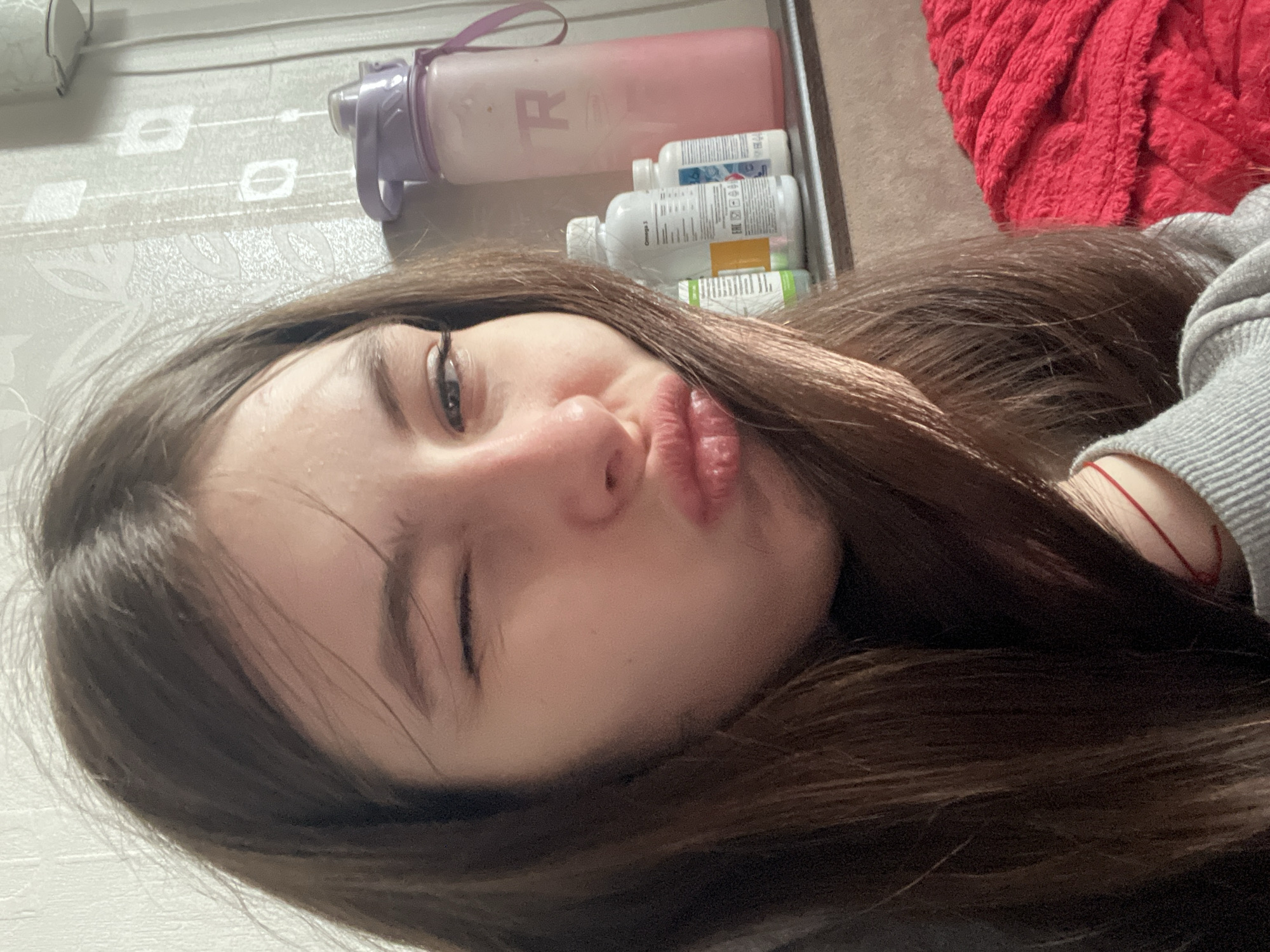 A close-up selfie, taken indoors, shows a young woman with long, brown hair. She is lying down and facing the camera, with one eye closed in a wink. Her lips are pursed. In the background, there are several objects on what appears to be a shelf: a pink water bottle, several white bottles with labels, and a red blanket. The wall behind the shelf is white with a subtle pattern. No specific details indicate the location in Saint Petersburg, Russia.