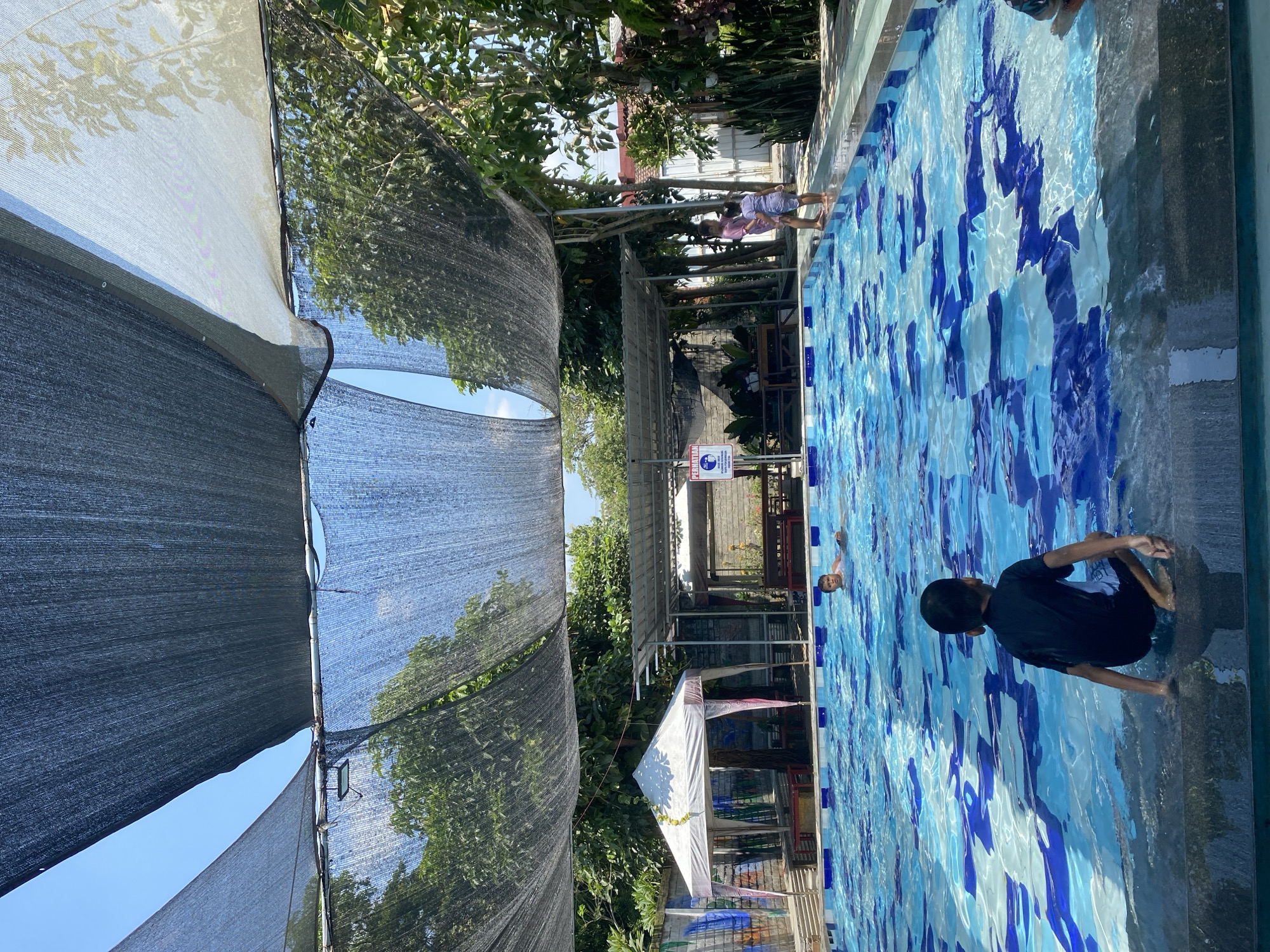 The image depicts an outdoor swimming pool facility, likely in Kediri City, Indonesia, on a sunny day. A boy in a black shirt and white shorts is sitting at the edge of the pool, facing away from the camera. In the background, another boy is swimming in the pool. Two young girls are standing by the pool's edge on the right side of the frame. The pool has a blue and white patterned bottom. Overhead, dark netting is stretched across poles to provide shade, with glimpses of the blue sky and green trees visible through the netting. Several structures are visible in the background, including a white tent with red trim, covered seating areas with metal roofs, and a brick wall with graffiti. A sign with "PERHATIAN" (Attention) and safety icons is posted on one of the structures. The overall environment appears to be a recreational area designed for swimming and relaxation.