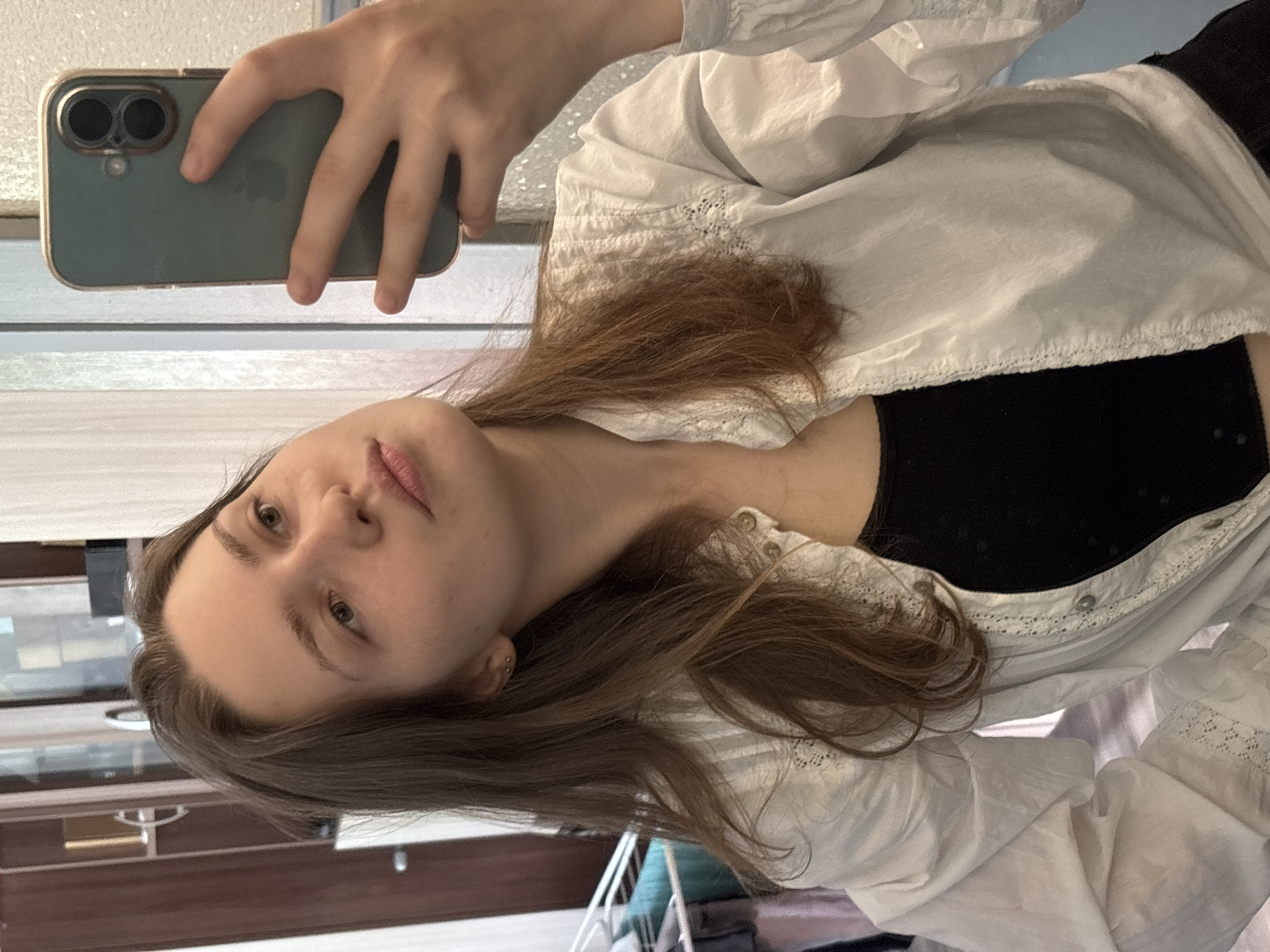 A person with fair skin and light brown hair takes a selfie with a green iPhone. The person is wearing a black top and a white, long-sleeved shirt with lace trim and buttons. The background shows what appears to be part of an interior with a window. The location cannot be definitively determined as Saint Petersburg, Russia based solely on the provided image.