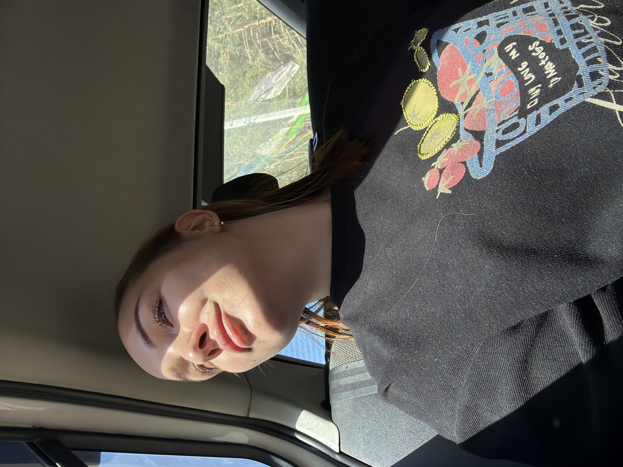 A young woman is inside a vehicle, likely a car or van, as evidenced by the interior panels and window. She is wearing a black sweatshirt with a colorful, stylized design on the front. Sunlight illuminates her face. The scene outside the window appears to be a wooded area. The image could have been taken in a vehicle traveling through or near Chaikovsky, Russia.