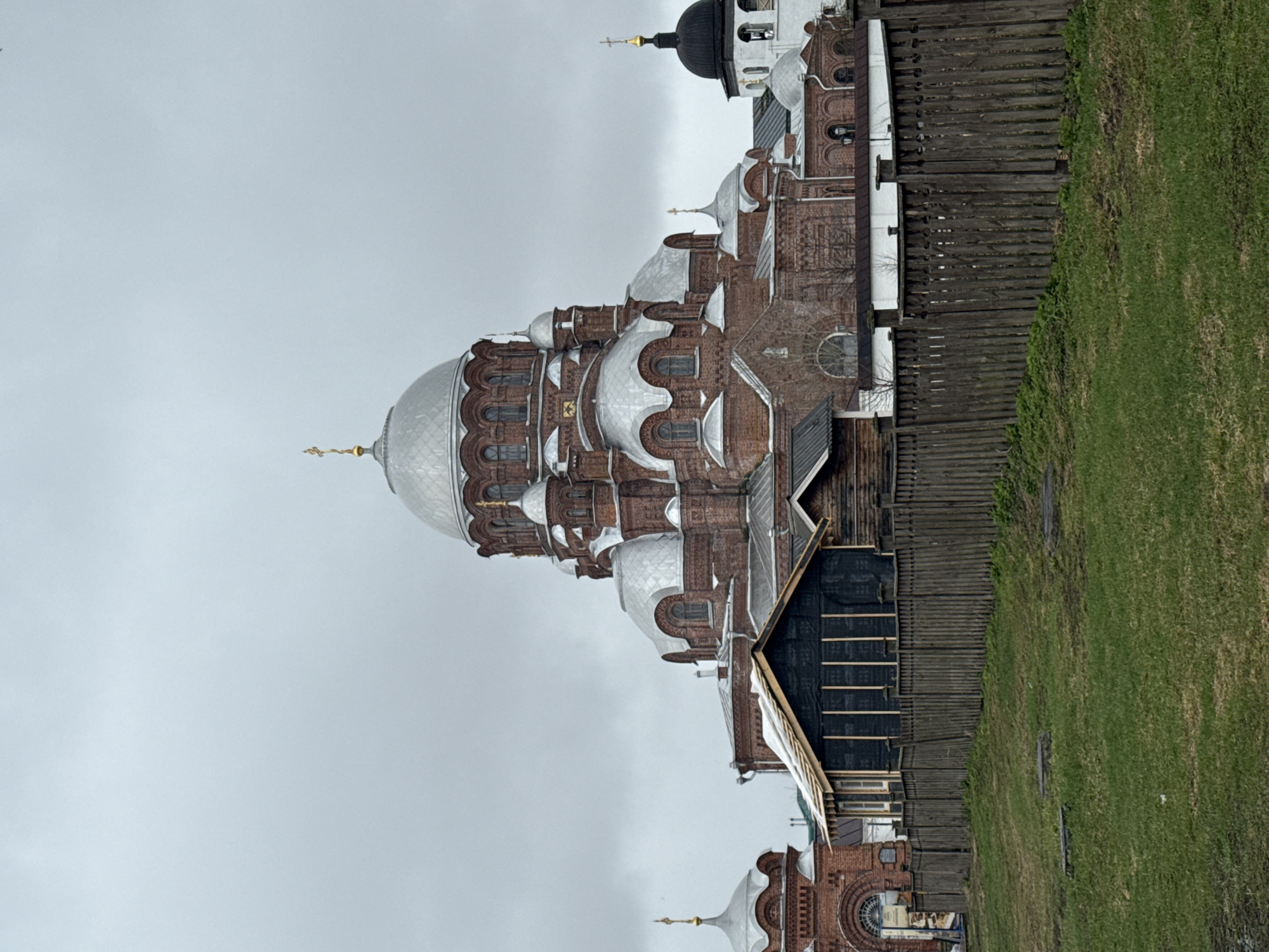 The image shows the Cathedral of the Icon of the Mother of God of All Who Sorrow in the city of Sviyazhsk, Russia. The cathedral features distinctive onion domes covered in silver-colored metal. The building is primarily constructed of red brick and includes smaller windows and architectural details. A wooden fence borders the church grounds, and a patch of green grass is visible. The sky overhead is overcast.
