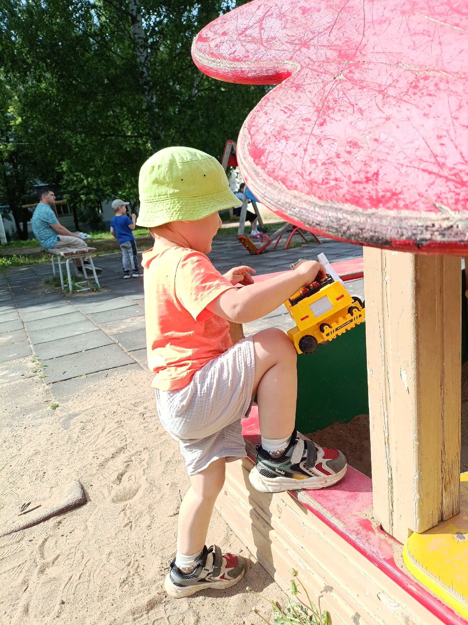 A young child wearing a lime green bucket hat, an orange t-shirt, and grey shorts is standing on the edge of a red and yellow playground structure. He is holding a yellow toy truck in his hands. The structure is surrounded by sand.
In the background, two men are visible on a paved area near trees. One man is sitting on a stool and the other is standing.
The location is the city of Chaikovsky, Russia.