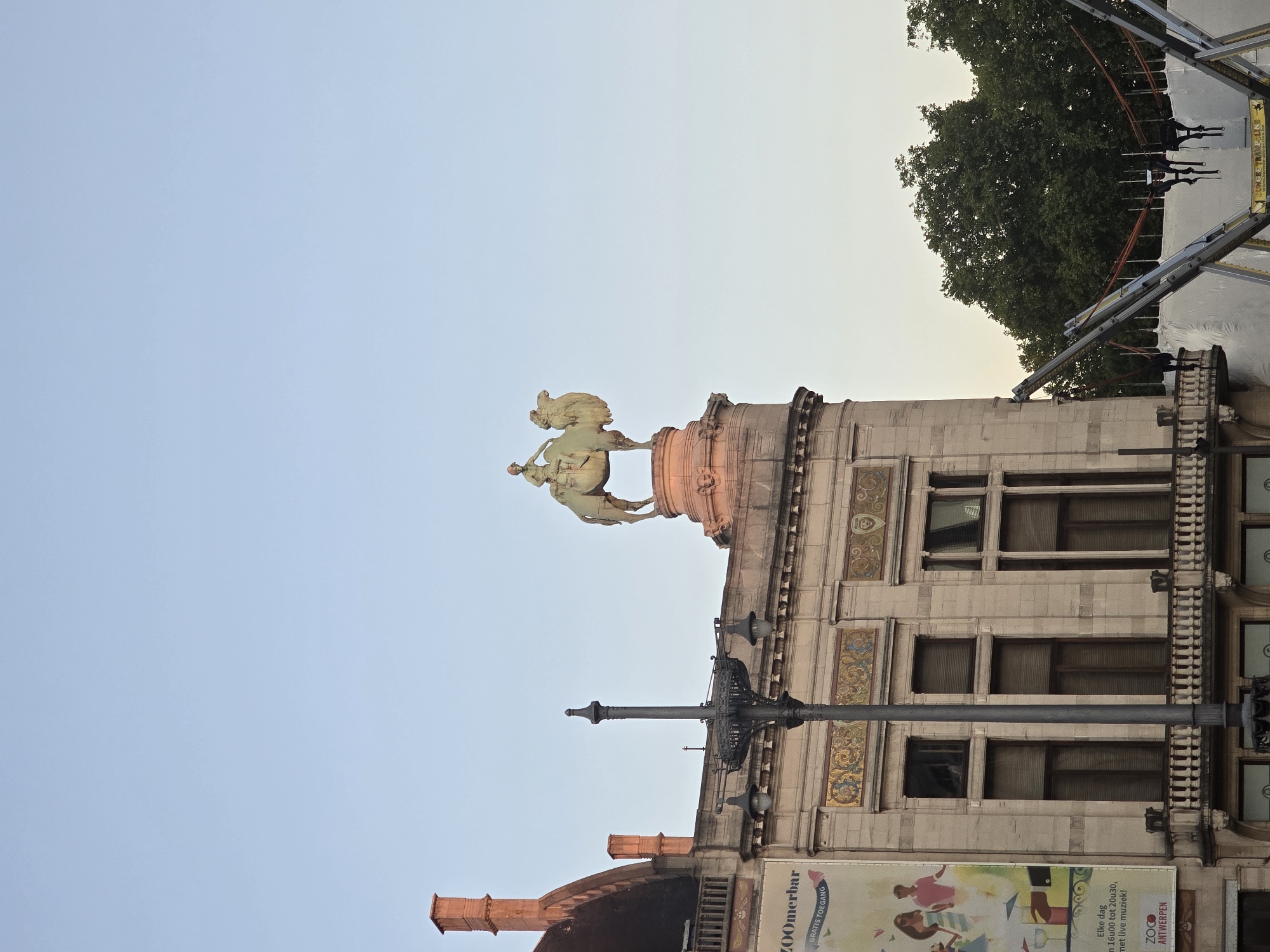 The image shows the facade of a building in Antwerp, Belgium, with a statue of a person riding a camel on top. The building appears to be old and ornate, with decorative carvings and columns. There is a banner on the left side of the building that says "ZOOMerbar GRATIS TOEGANG" and mentions "live muziek". A lamppost stands in front of the building. In the background, there are trees and a blue sky. The lower right of the image shows part of a bungee jumping attraction. The overall impression is of a historical building with a modern attraction nearby, possibly in a park or public square.