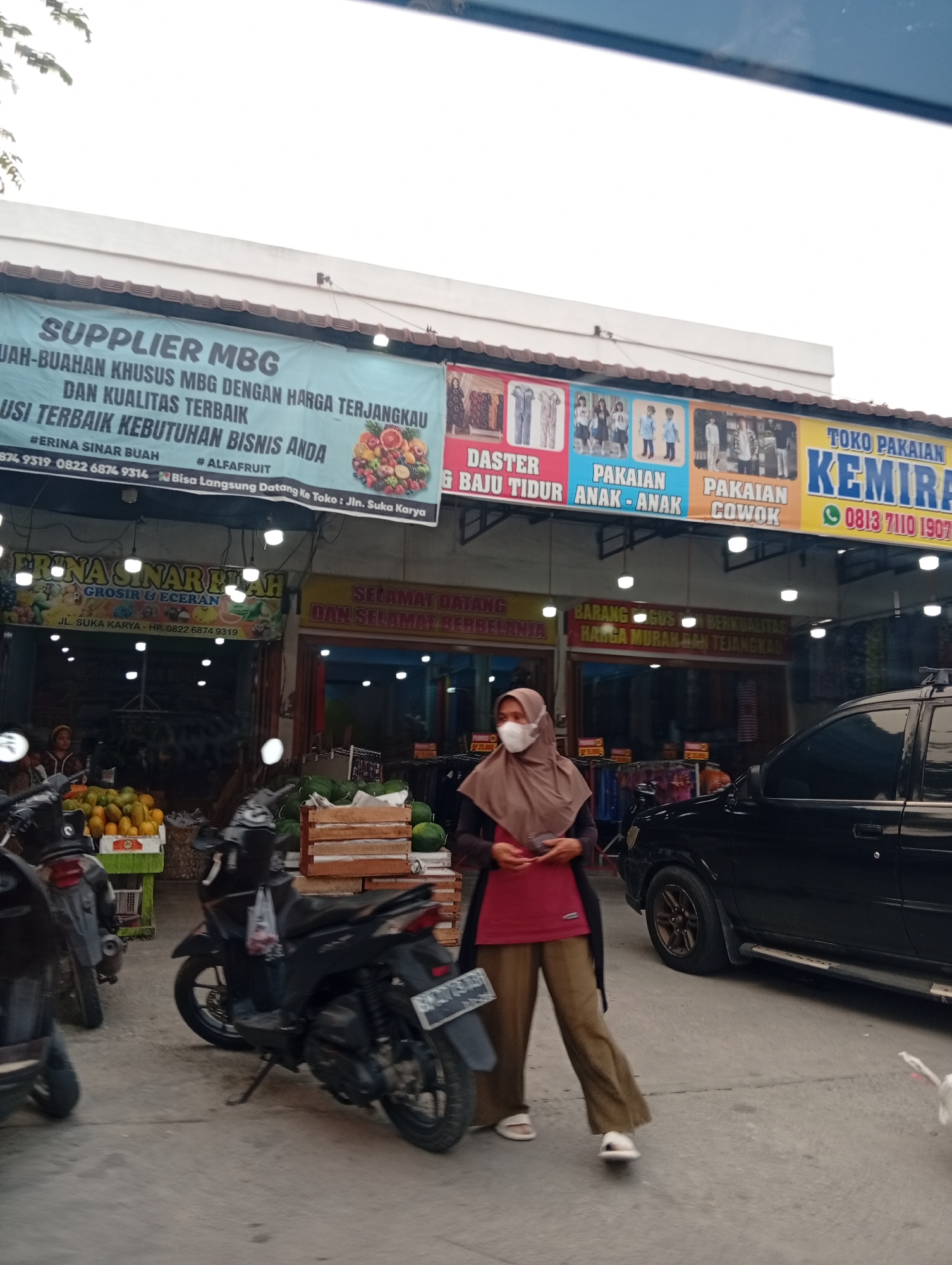 A street-level view captures a busy commercial area in Tarai Bangun, Indonesia, with several shops and stalls. In the foreground, a person wearing a hijab and a face mask walks past a black SUV, with two motorbikes parked to the left. Behind them, a fruit stall displays a variety of colorful fruits, with crates of produce stacked nearby. The shops feature prominent signage, including "SUPPLIER MBG BUAH-BUAHAN KHUSUS MBG DENGAN HARGA TERJANGKAU DAN KUALITAS TERBAIK" (Supplier MBG Special Fruits at Affordable Prices and Best Quality) and "TOKO PAKAIAN KEMIRA" (Kemira Clothing Store), indicating a mix of businesses catering to food and apparel. A banner above the shops advertises clothing items like "DASTER BAJU TIDUR" (Daster Sleepwear), "PAKAIAN ANAK-ANAK" (Children's Clothing), and "PAKAIAN COWOK" (Men's Clothing). The environment appears to be an outdoor marketplace, with goods displayed openly and shoppers moving through the area. The overall impression is of a vibrant, active commercial hub in Indonesia.