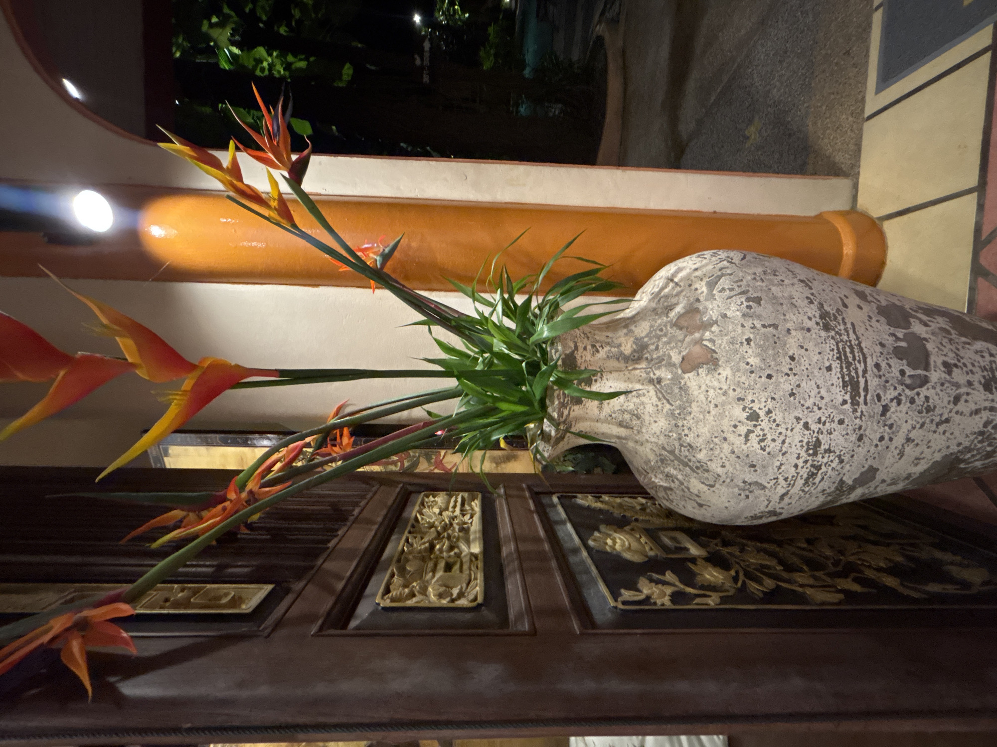 The image shows a large, distressed ceramic vase filled with a bouquet of vibrant orange and yellow heliconia flowers and green foliage. The vase is positioned next to a dark wooden door with intricate gold relief carvings. To the right of the vase, a thick, orange-painted pillar with a white trim supports an arched doorway, through which a dimly lit outdoor area with trees and possibly a pool is visible. A bright white spotlight is mounted on the pillar, illuminating the area. The flooring consists of square tiles in various earthy tones. The scene appears to be an entryway or decorative arrangement, possibly at a resort or upscale establishment in Chalong, Thailand, given the tropical flora and architectural style. There are no people or discernible actions taking place.