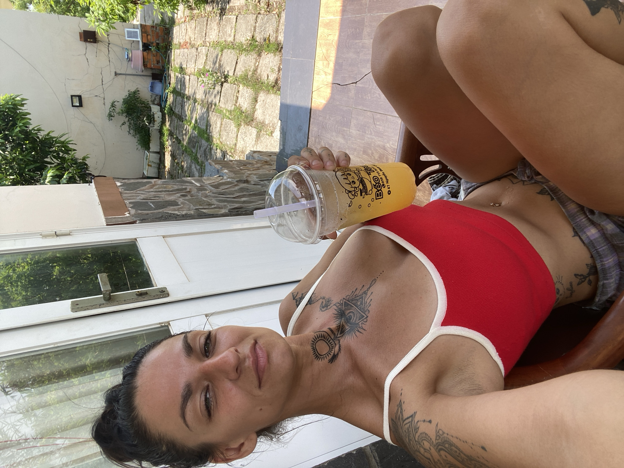 A woman with dark hair pulled up in a bun and tattoos on her neck, chest, and arms is sitting outside on a chair, holding a plastic cup of a yellow beverage with a straw. She is wearing a red tank top and plaid shorts. The background shows a patio area with stone tiles and some greenery. A white door with a metal handle is visible to her left, and part of a building with a stone facade is to her right. The scene appears to be in Mũi Né, Vietnam, suggested by the architecture and the text on the beverage cup.