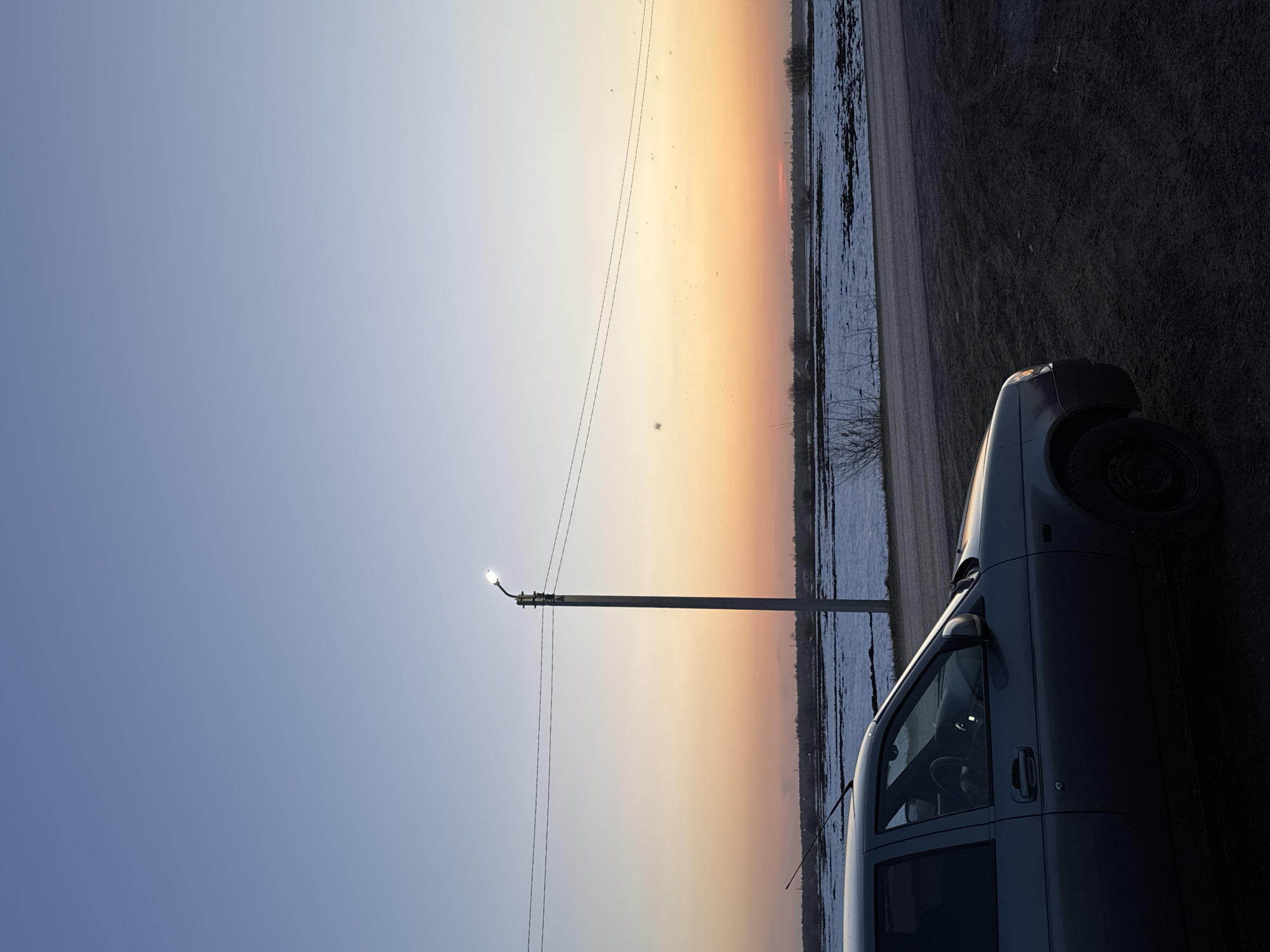 The image shows a sunset scene in or near the city of Кондратово, Russia. The sky transitions from blue to orange above a landscape with snow-covered fields. A street lamp is visible on the left with wires extending across the frame. A gray car is parked on the right side of the image.