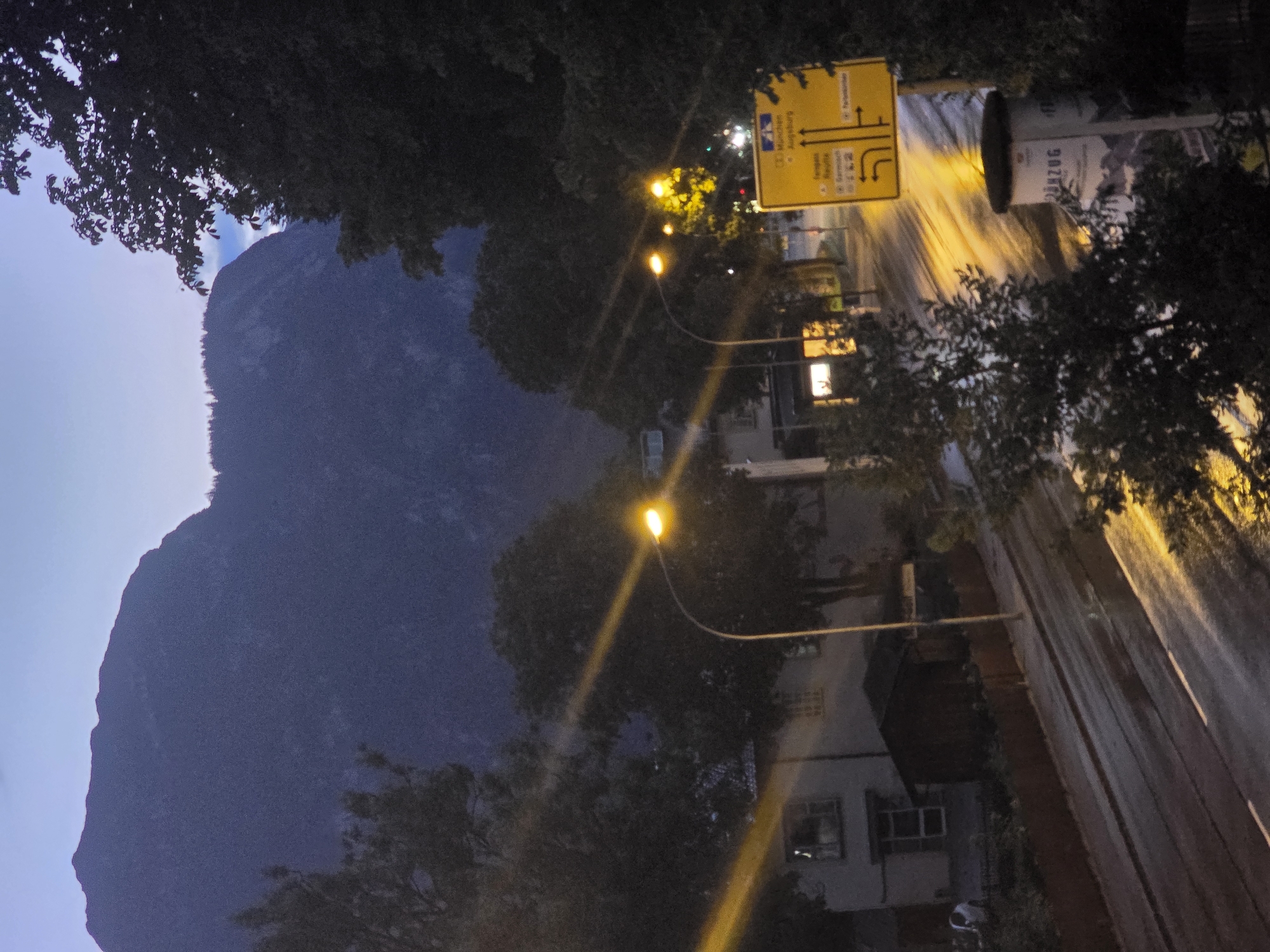 The image depicts a street scene in Garmisch-Partenkirchen, Germany, during dusk. A large mountain range dominates the background, its silhouette dark against the twilight sky. Trees flank the street, some with illuminated streetlights casting warm glows. On the right side of the image, a yellow directional signpost indicates routes to München and Augsburg, with smaller towns such as Tegernsee and Garmisch listed. The street appears wet, suggesting recent rain, with reflections of the streetlights on the surface. Buildings line the left side of the street, some with illuminated windows. A small shed is visible in front of one of the buildings. A cylindrical bin with German text, possibly advertising a tourist attraction like a cogwheel train ("ZÜG"), stands near the signpost. No individuals are clearly discernible in the image.