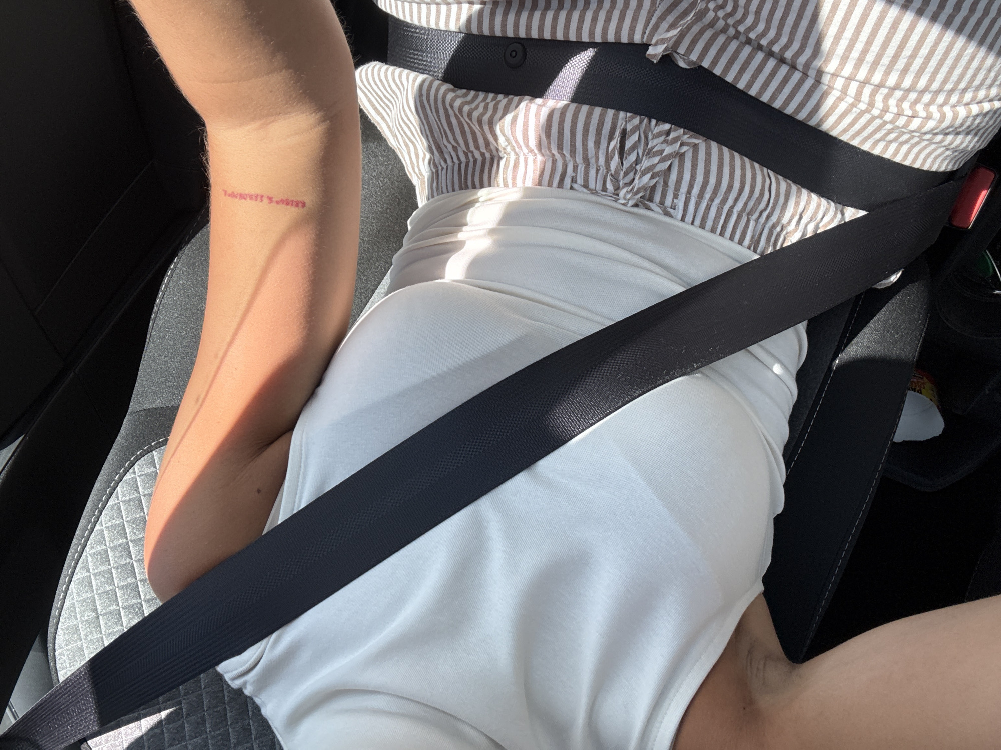 The image depicts a person wearing a white tank top and a striped shirt in a car, fastened with a seatbelt. The person has a tattoo on their left arm that reads "LOCATION'S NAME". Based on the tattoo and the context of the prompt, it's possible the location refers to Le Muy, France. The person is sitting on a gray car seat.
