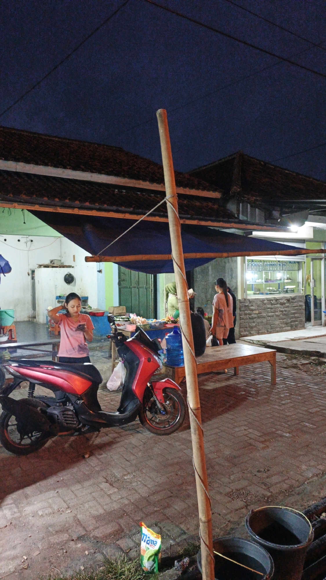 In Cibenda, Indonesia, at dusk, a roadside scene unfolds. A red scooter is parked on a brick-patterned ground in front of a stall. A young woman, wearing a pink t-shirt and black shorts, stands beside the scooter, looking at her phone. Behind her, a blue tarp is stretched over the stall. Another woman in a green hijab is visible in the background. To the right, a wooden bench is placed next to the stall, and two large black buckets are near a drain. The sky is dark blue, suggesting the night has begun. A tall bamboo pole stands in the foreground, tied with rope to the tarp. A packet of Mama brand seasoning is on the ground near the scooter.