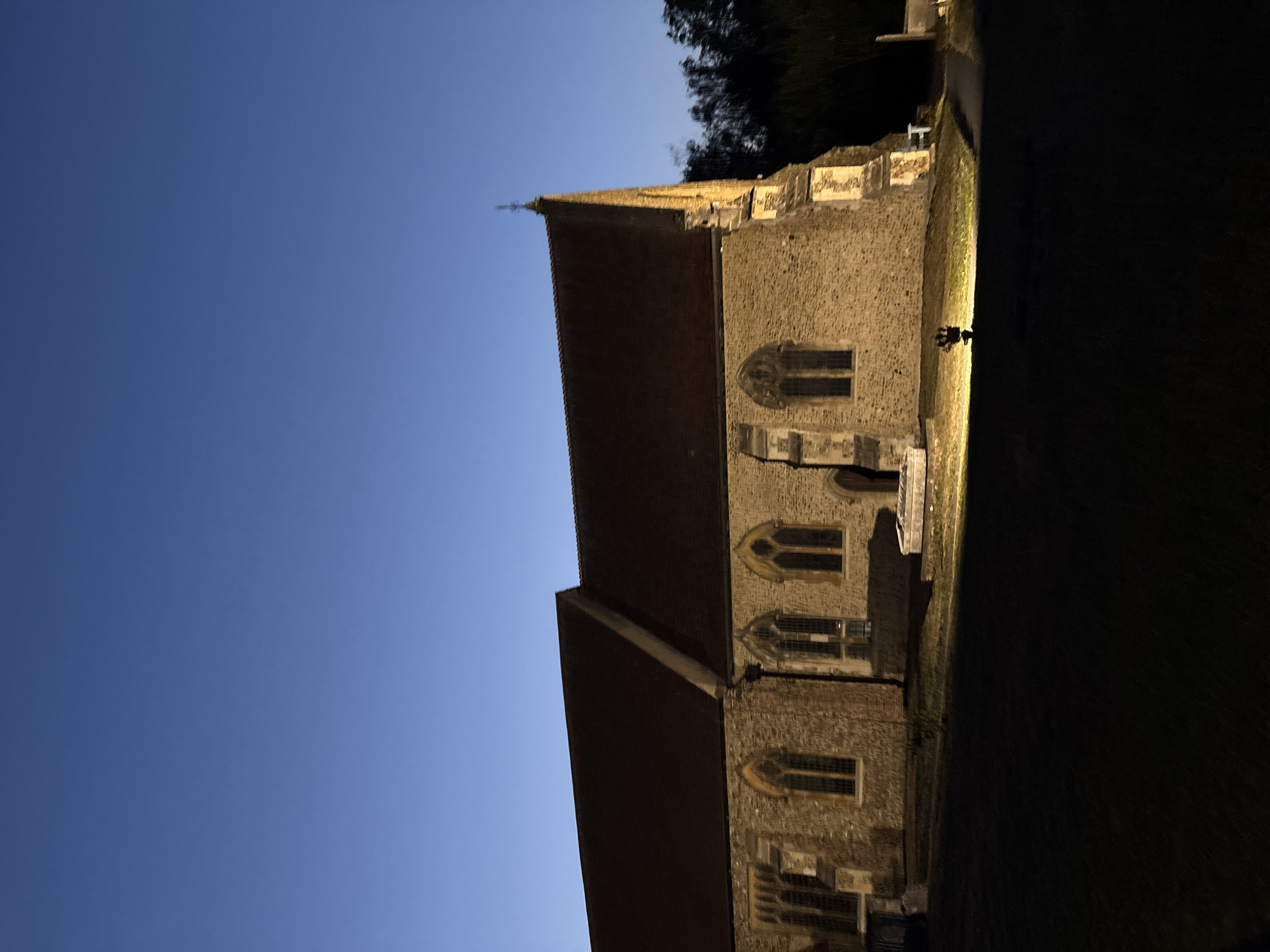 An exterior, low-angle, wide shot captures a stone church building illuminated by exterior lighting at dusk. The sky is a deep, clear blue transitioning to darker shades towards the horizon. The church, constructed of light-colored stone with a dark brown, tiled roof, features arched windows with gothic detailing. A cross tops the highest point of the roof. The foreground consists of dark, shadowed grass and a paved pathway leading away from the building. The scene is devoid of any people or animals. The architecture suggests a historical building, possibly a parish church, given its style and presence within a grassy area that could be a churchyard. The lighting creates strong contrasts between the illuminated facade and the shadowed grounds.